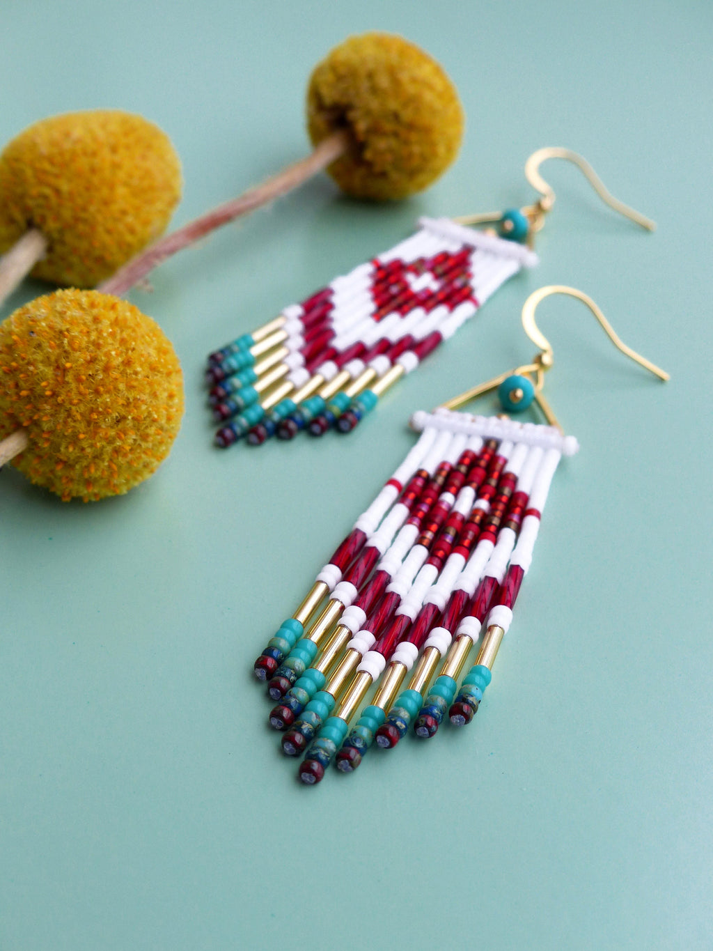 Peut inclure: Une paire de longues boucles d'oreilles en perles avec un motif géométrique en rouge, blanc et turquoise. Les boucles d'oreilles ont des accents dorés et pendent à des crochets dorés.