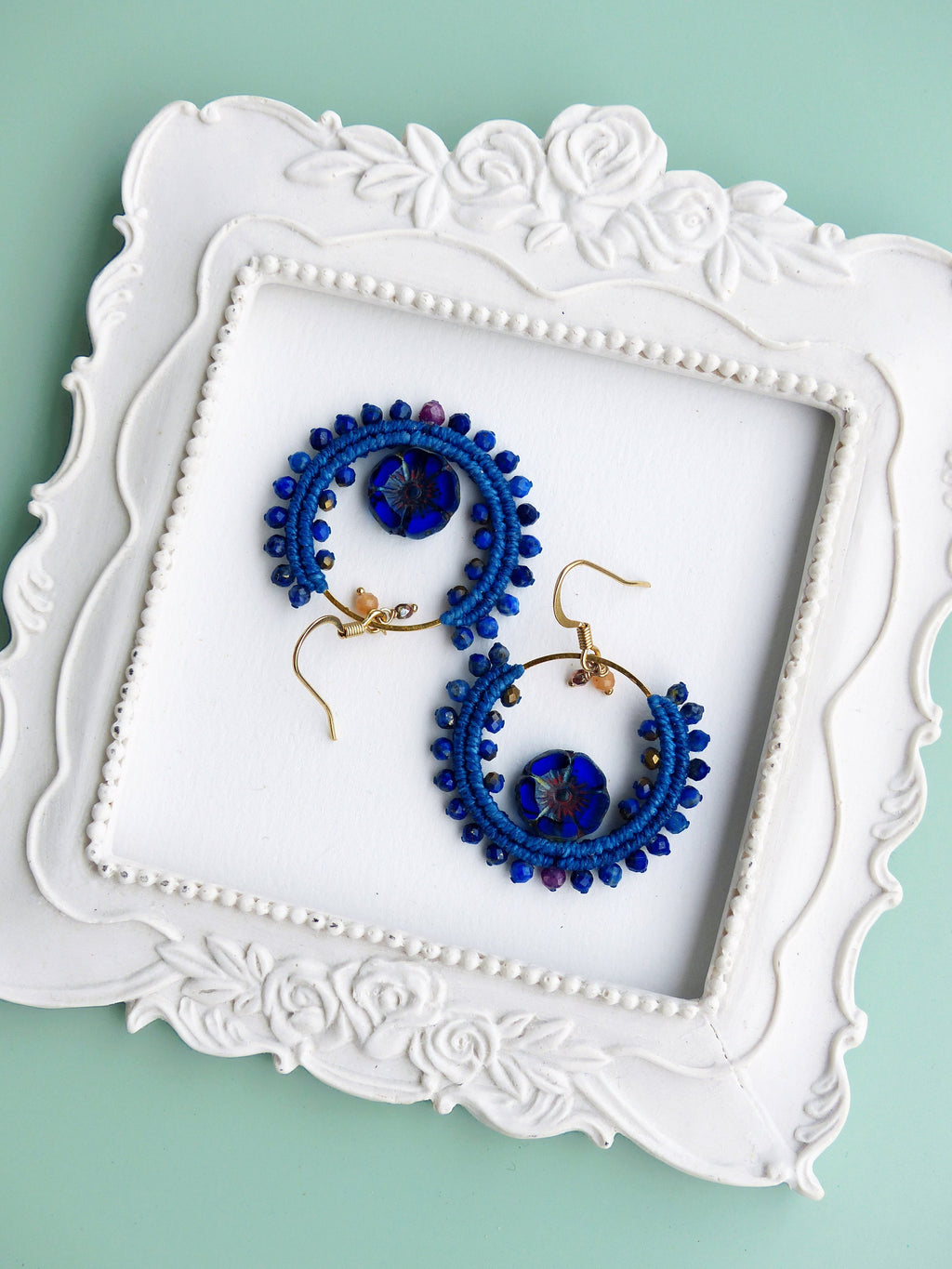 Peut inclure: Une paire de boucles d'oreilles créoles bleues et dorées avec un charme de fleur en verre bleu. Les boucles d'oreilles sont présentées sur un plateau blanc à motif floral.