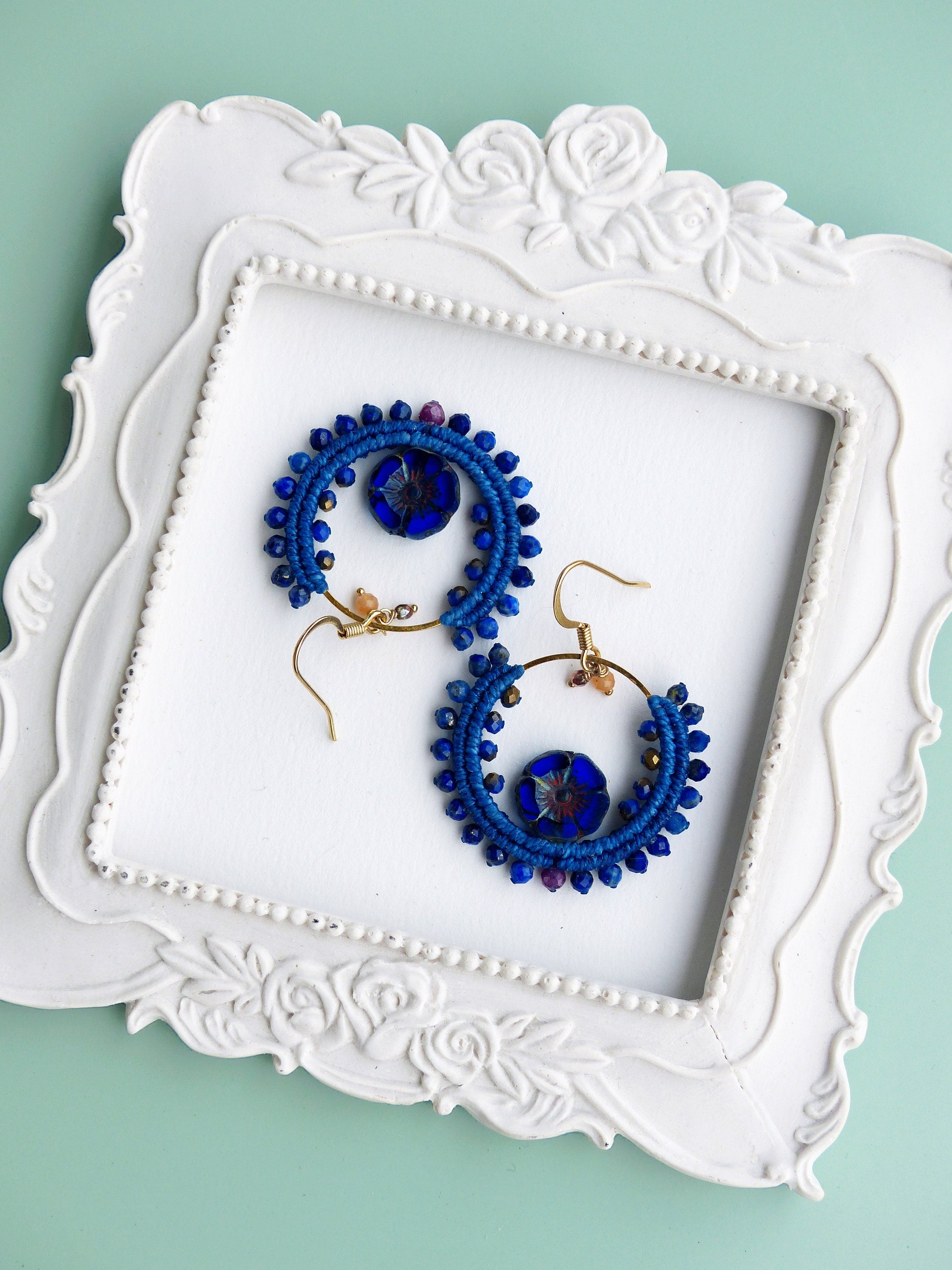 Peut inclure: Une paire de boucles d'oreilles créoles bleues et dorées avec un charme de fleur en verre bleu. Les boucles d'oreilles sont présentées sur un plateau blanc à motif floral.