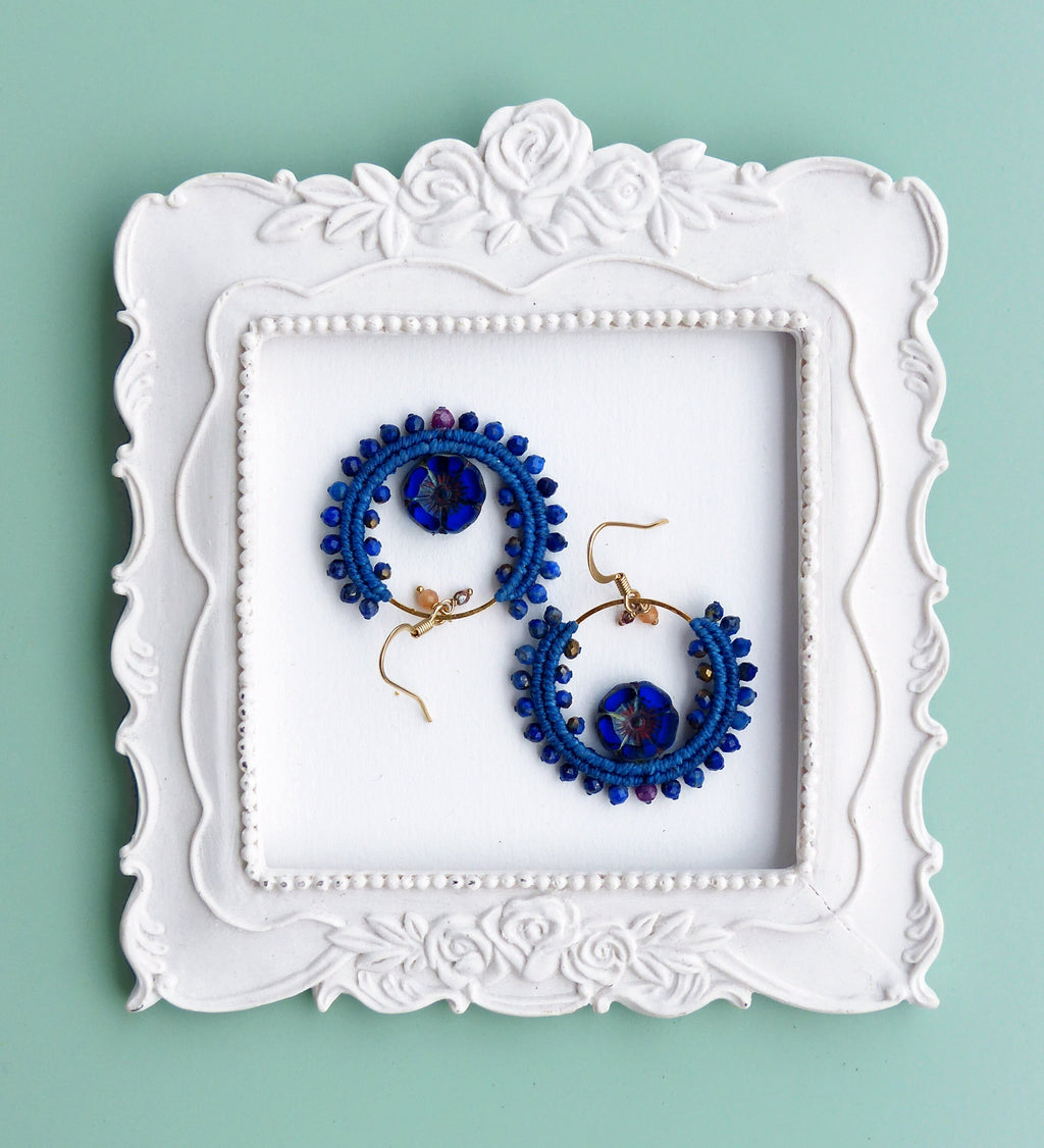 Peut inclure: Une paire de boucles d'oreilles créoles bleues et dorées avec un motif perlé. Les boucles d'oreilles sont présentées sur un cadre blanc à motif floral.