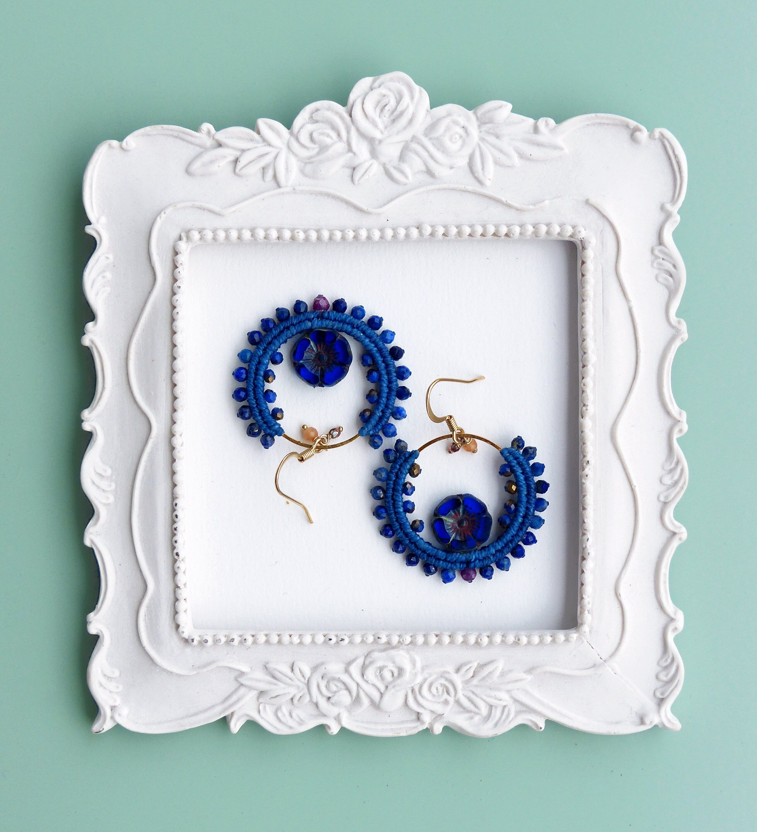 Peut inclure: Une paire de boucles d'oreilles créoles bleues et dorées avec un motif perlé. Les boucles d'oreilles sont présentées sur un cadre blanc à motif floral.