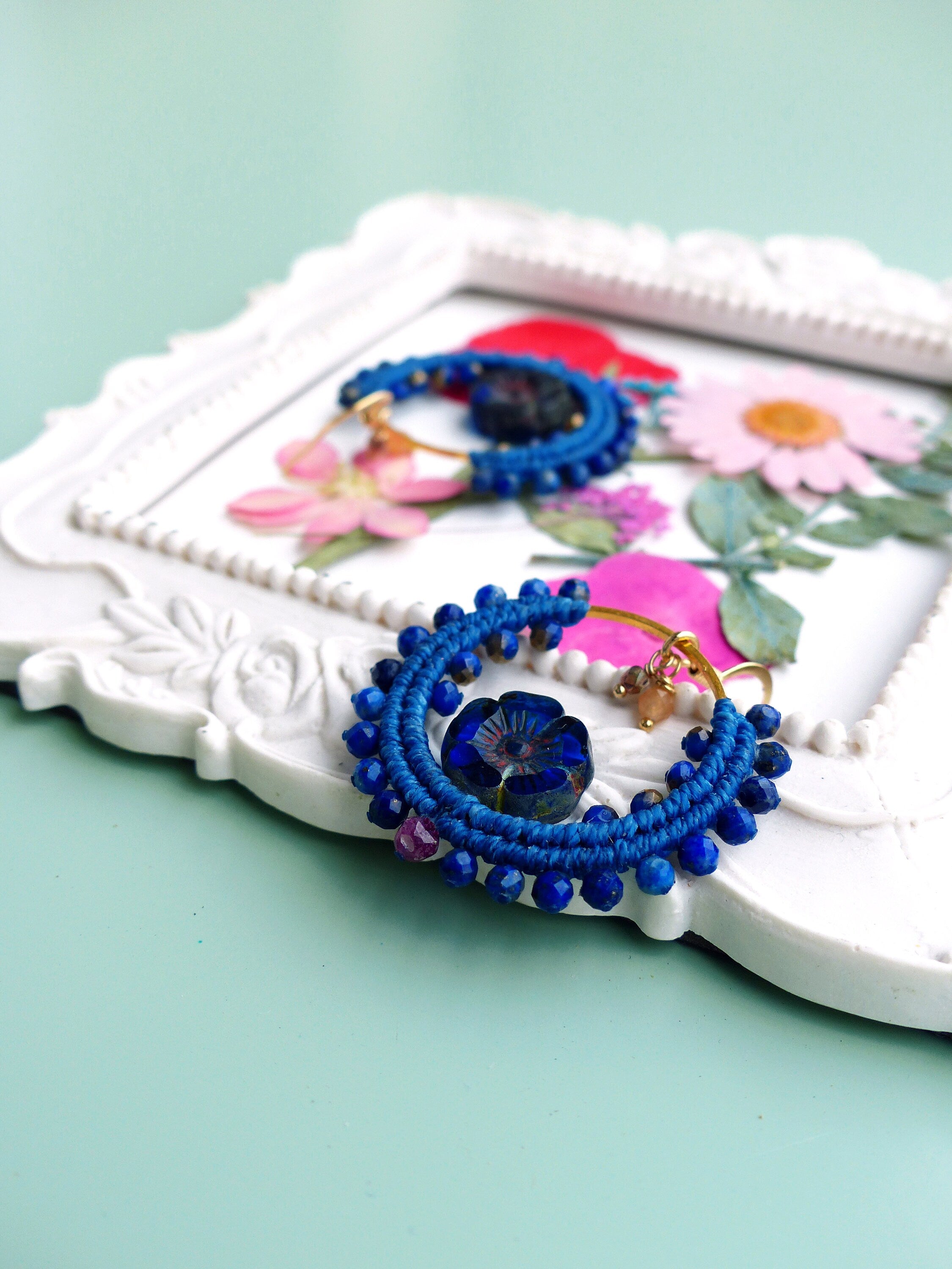 Peut inclure: Une paire de boucles d'oreilles créoles bleues et dorées avec un charme de fleur bleue. Les boucles d'oreilles sont faites de perles bleues et de fil d'or. Les boucles d'oreilles sont présentées sur un cadre blanc avec des fleurs séchées.