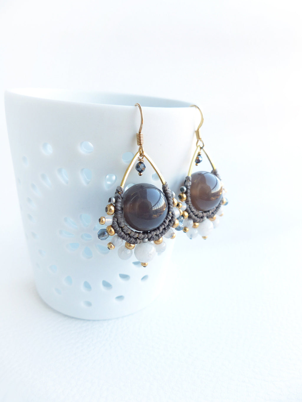 Peut inclure: Une paire de boucles d'oreilles artisanales avec de grosses perles de pierres précieuses gris fumé. Les boucles d'oreilles ont une forme de goutte d'eau avec des montures en fil d'or, du macramé gris et des accents de petites perles dorées, blanches et grises.