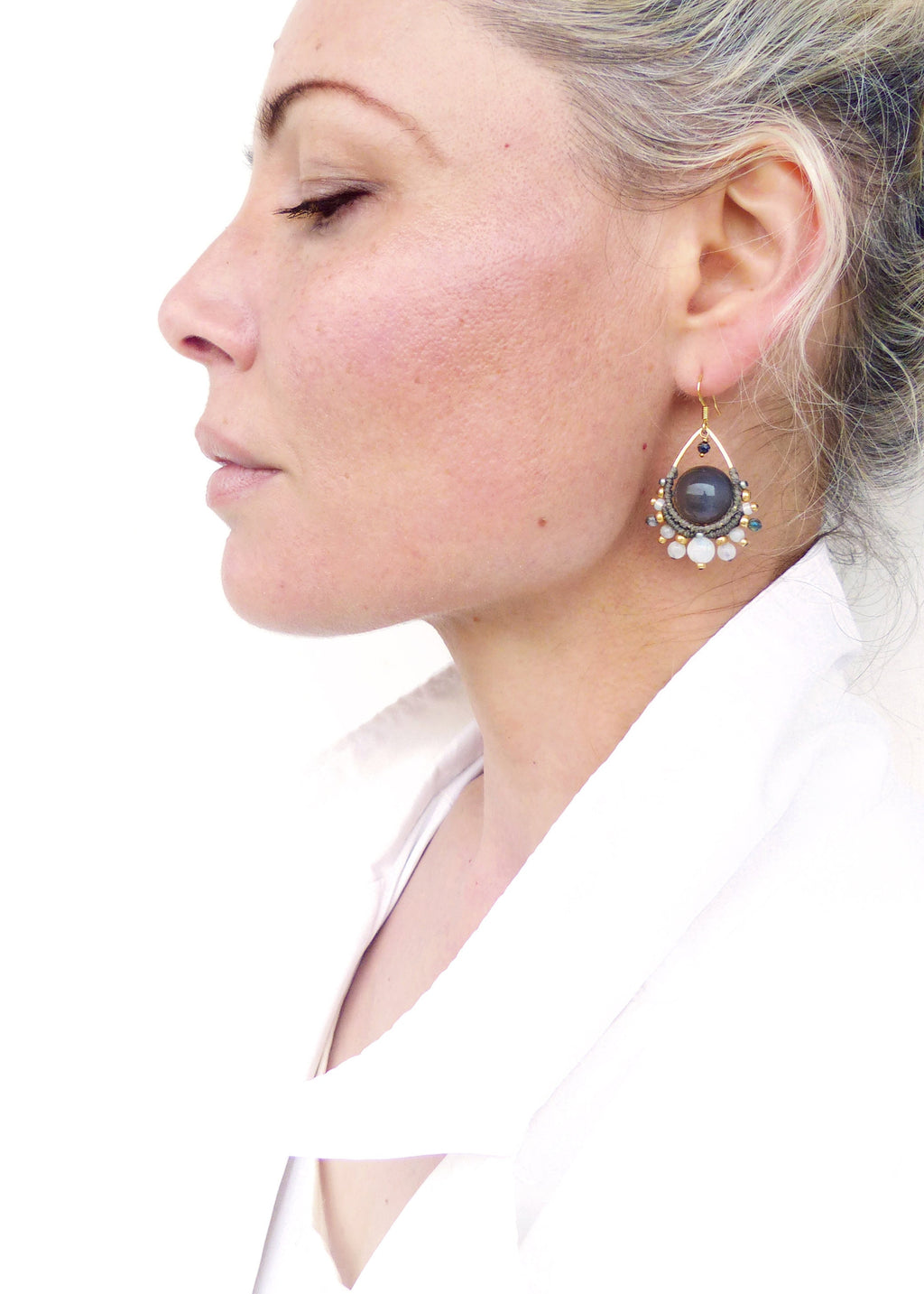 Peut inclure: Gros plan d'une femme portant une boucle d'oreille dorée. La boucle d'oreille présente une grosse pierre gris foncé au centre, entourée de petites perles et pierres claires. La boucle d'oreille a une forme de goutte d'eau et un crochet doré.
