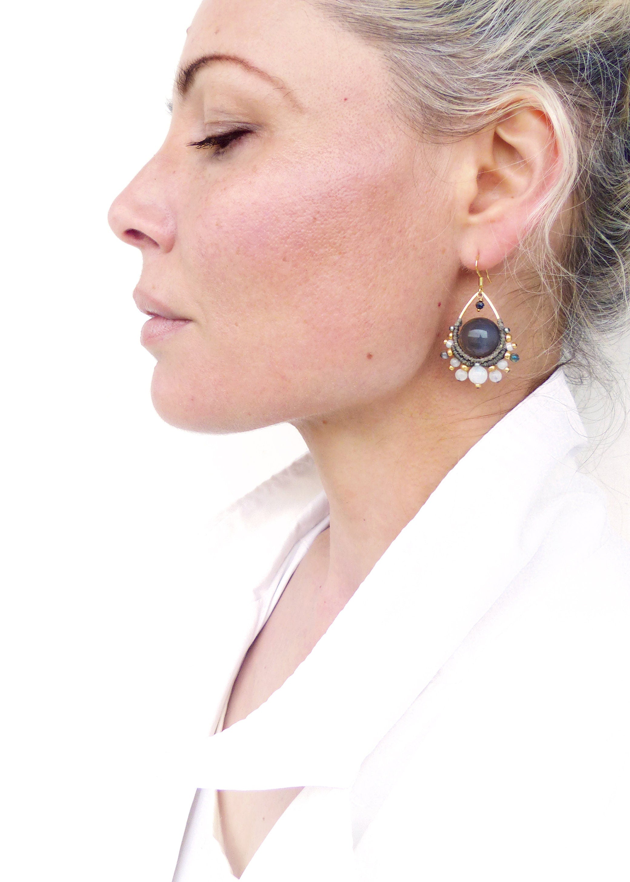 Peut inclure: Gros plan d'une femme portant une boucle d'oreille dorée. La boucle d'oreille présente une grosse pierre gris foncé au centre, entourée de petites perles et pierres claires. La boucle d'oreille a une forme de goutte d'eau et un crochet doré.