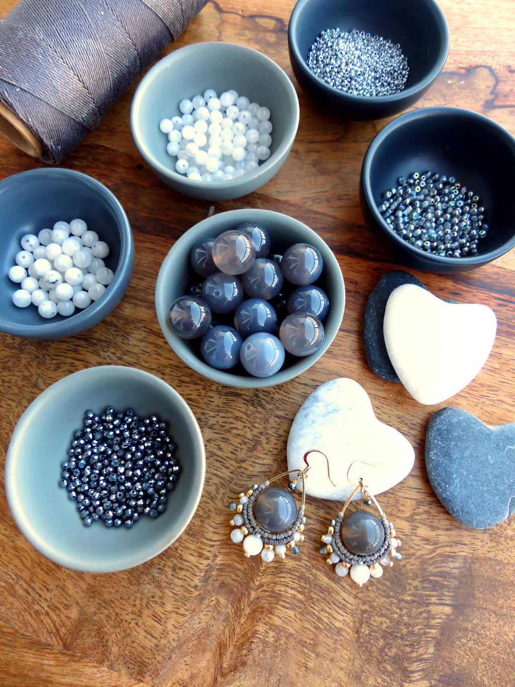 Peut inclure: Un assortiment de fournitures pour la création de bijoux, comprenant de petits bols gris remplis de perles de différentes tailles et couleurs, ainsi qu'une bobine de fil gris et une paire de boucles d'oreilles artisanales. Les boucles d'oreilles présentent de grosses perles grises et des accents dorés.