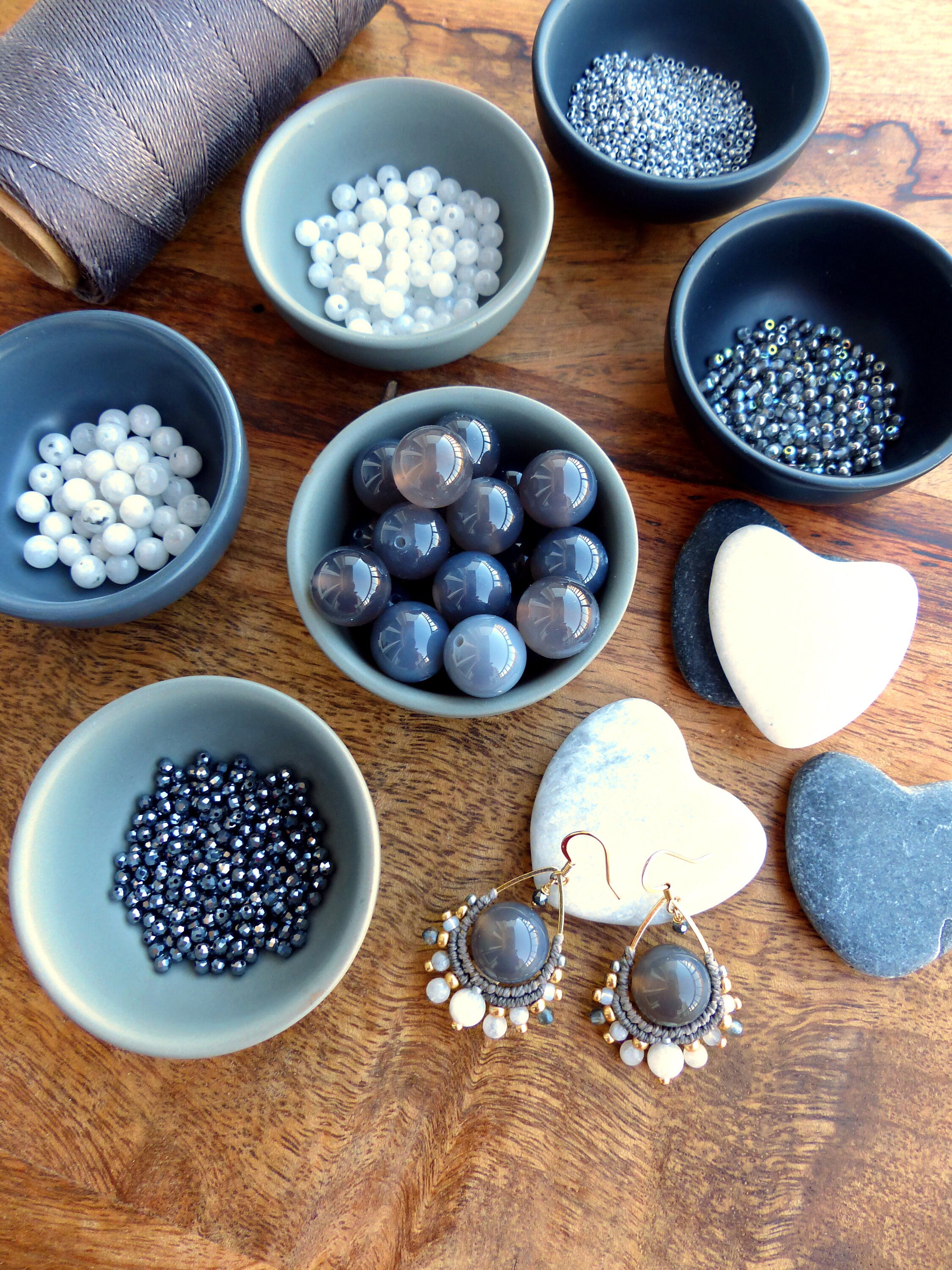 Peut inclure: Un assortiment de fournitures pour la création de bijoux, comprenant de petits bols gris remplis de perles de différentes tailles et couleurs, ainsi qu'une bobine de fil gris et une paire de boucles d'oreilles artisanales. Les boucles d'oreilles présentent de grosses perles grises et des accents dorés.