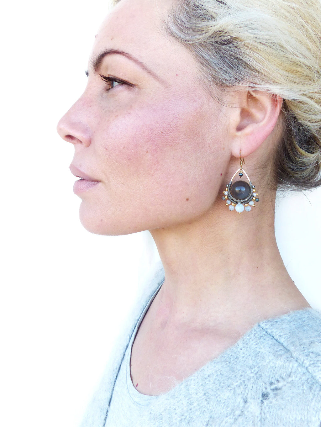 Peut inclure: Boucle d'oreille dorée en forme de goutte d'eau, ornée d'une grosse pierre gris foncé au centre, entourée de petites perles blanches et grises. La boucle d'oreille est portée à l'oreille, avec un pull bleu clair en arrière-plan.
