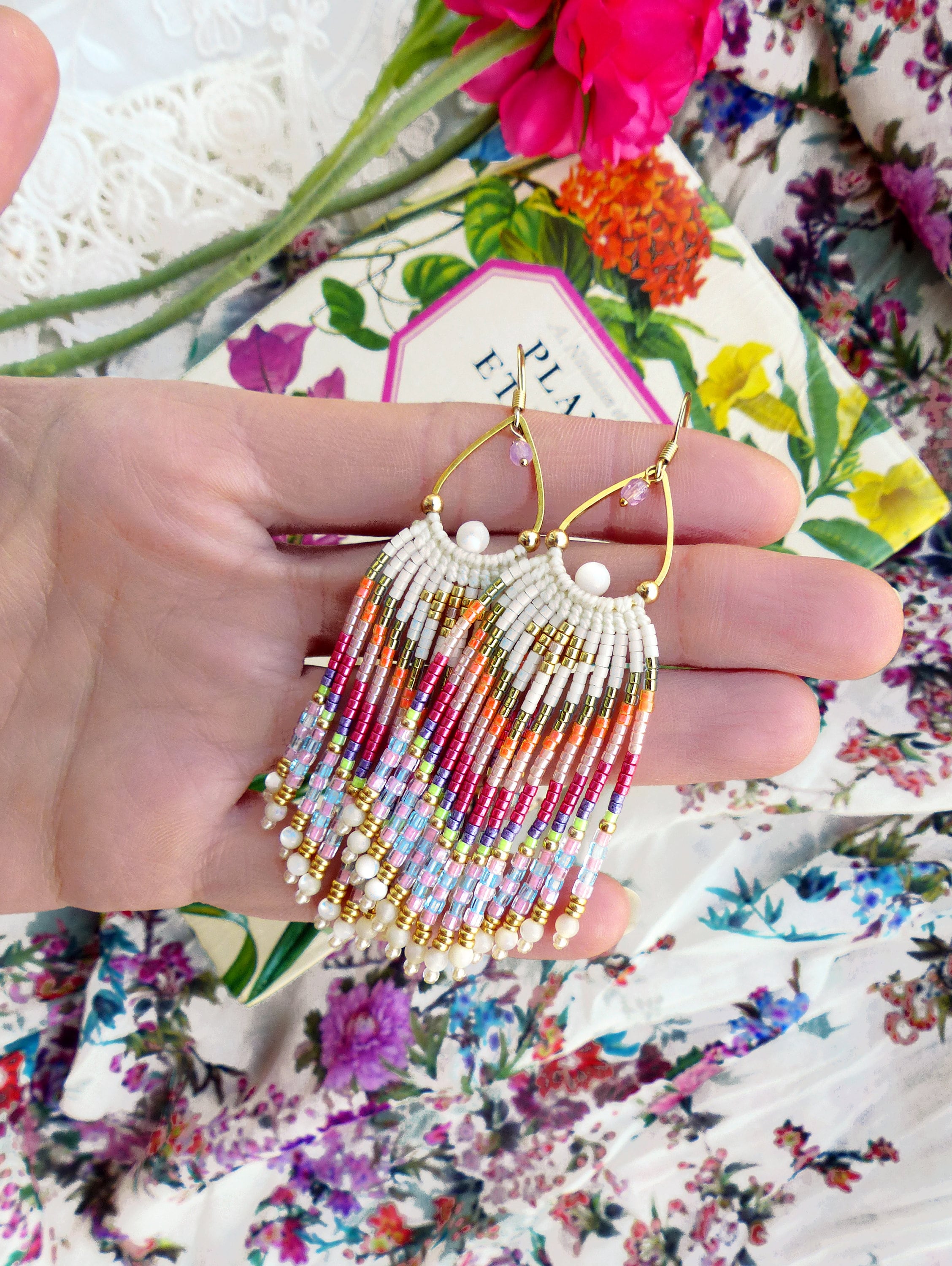 Peut inclure: Une paire de longues boucles d'oreilles en perles colorées avec des accents dorés et une forme de larme. Les boucles d'oreilles sont faites avec une variété de perles, y compris des perles blanches, roses, orange, violettes et bleues. Les boucles d'oreilles sont suspendues à des crochets dorés.
