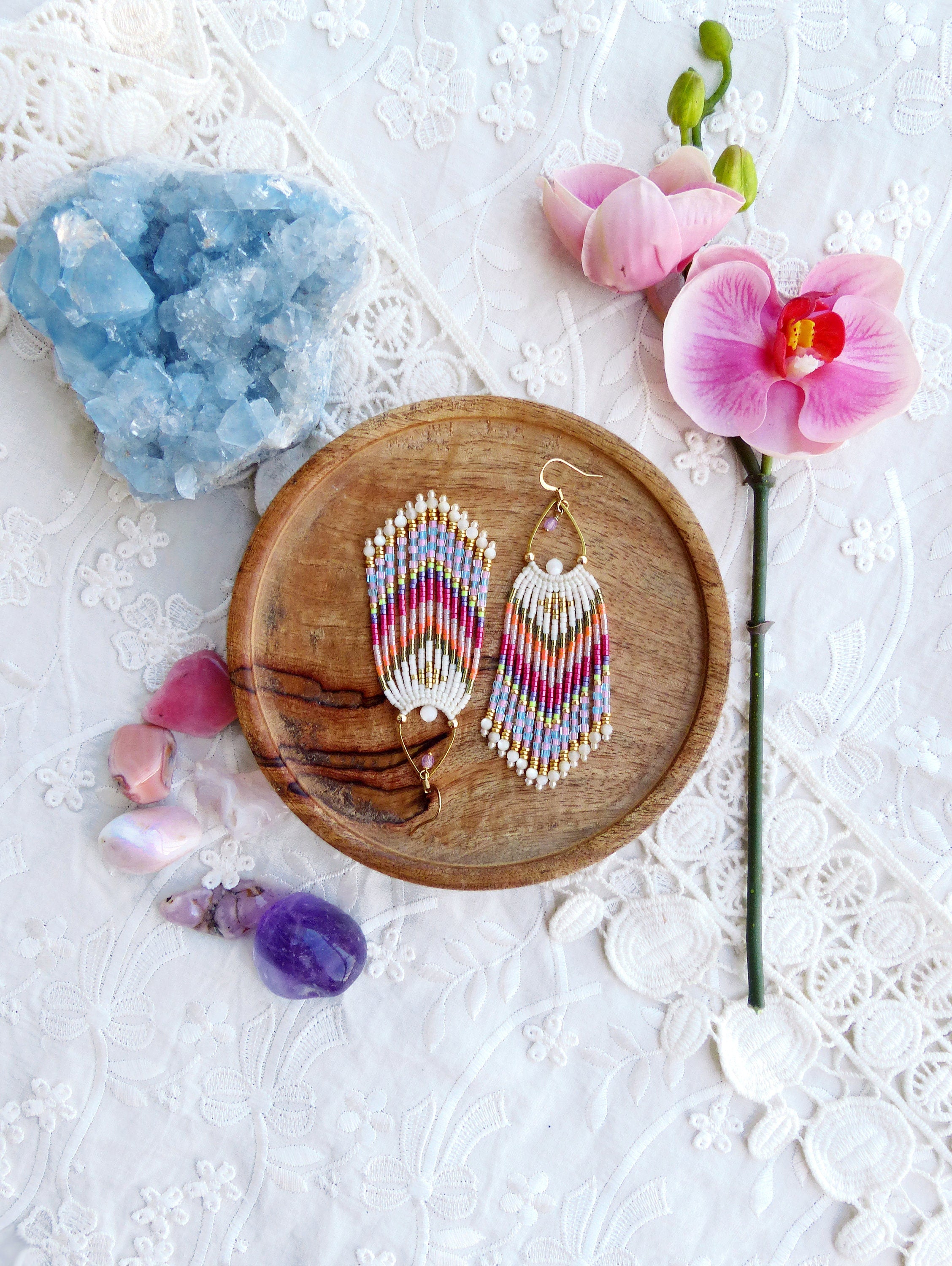 Peut inclure: Une paire de boucles d'oreilles en perles colorées avec une base blanche et des accents dorés. Les boucles d'oreilles sont présentées sur une assiette en bois avec un cristal bleu et une orchidée rose.