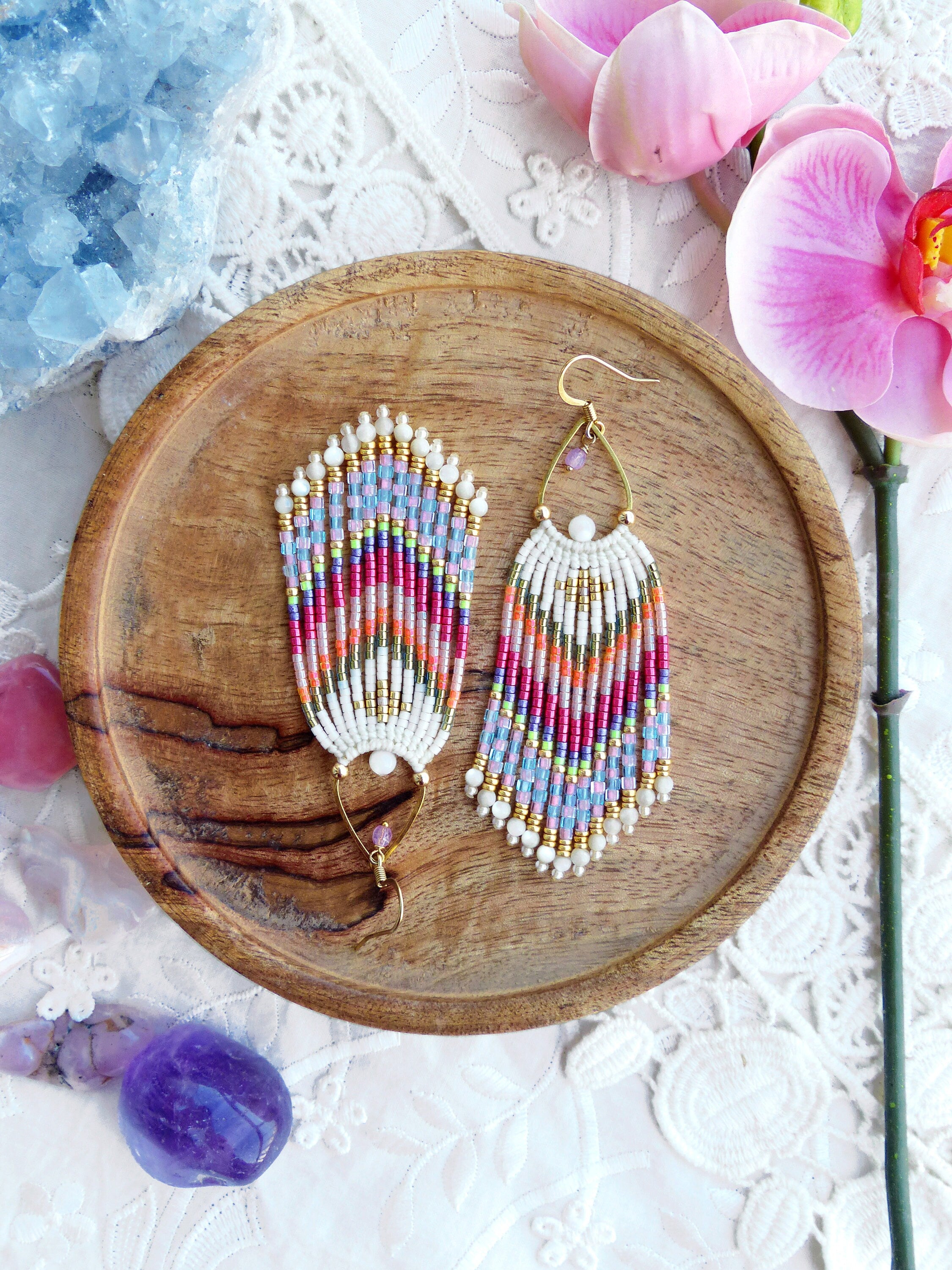 Peut inclure: Une paire de longues boucles d'oreilles en perles colorées avec des crochets dorés. Les boucles d'oreilles sont faites avec une variété de perles, y compris le blanc, le rose, le bleu, le vert et l'orange. Les boucles d'oreilles sont suspendues à une assiette en bois.