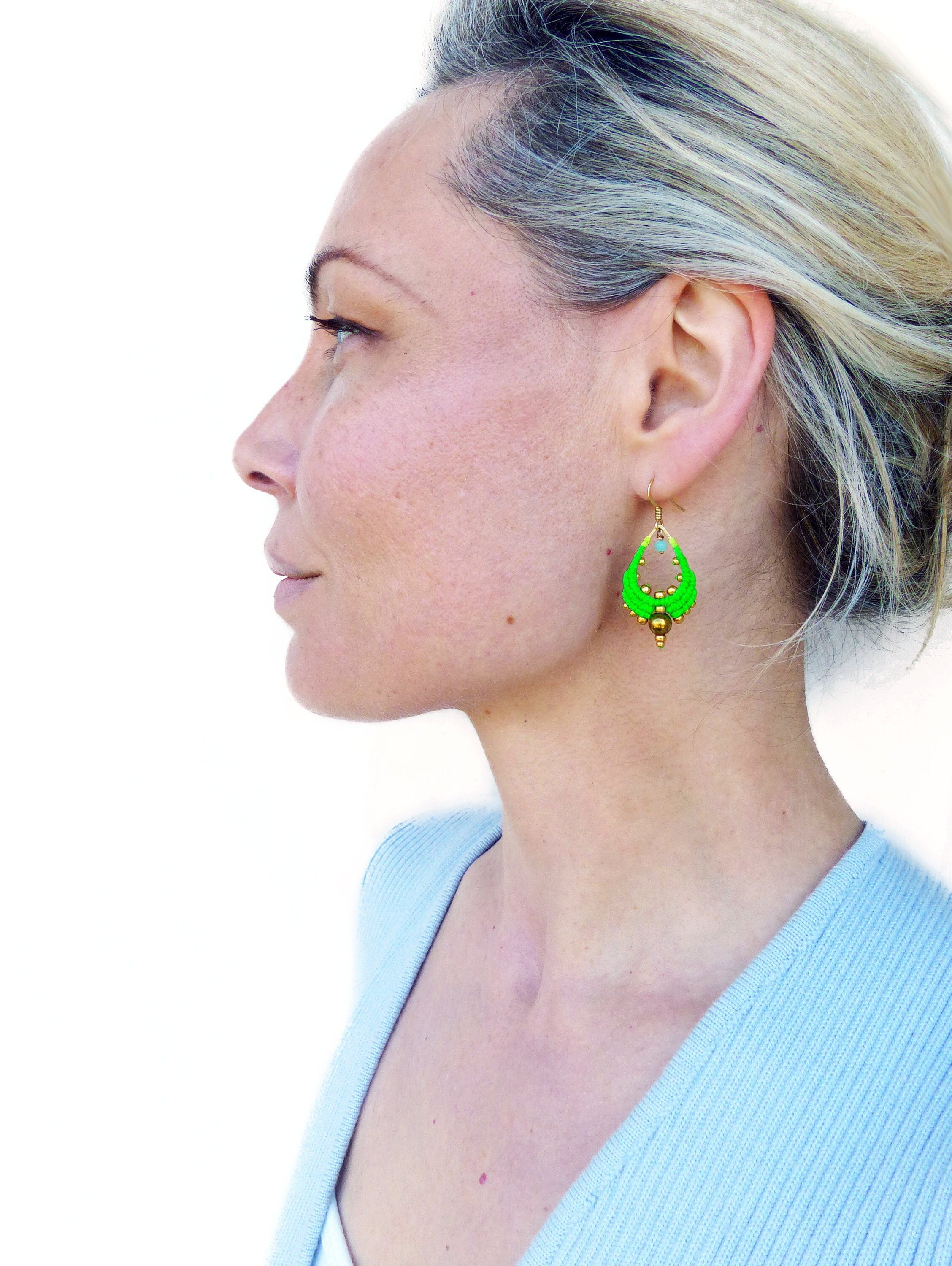 Peut inclure: Une femme portant un pull bleu clair avec des boucles d'oreilles pendantes vertes et dorées. Les boucles d'oreilles sont faites de petites perles et ont une forme de larme.