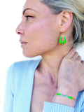 Peut inclure: Une femme portant un pull bleu clair avec un bracelet en perles vertes et dorées au poignet. Elle porte également une paire de boucles d'oreilles pendantes vertes et dorées.