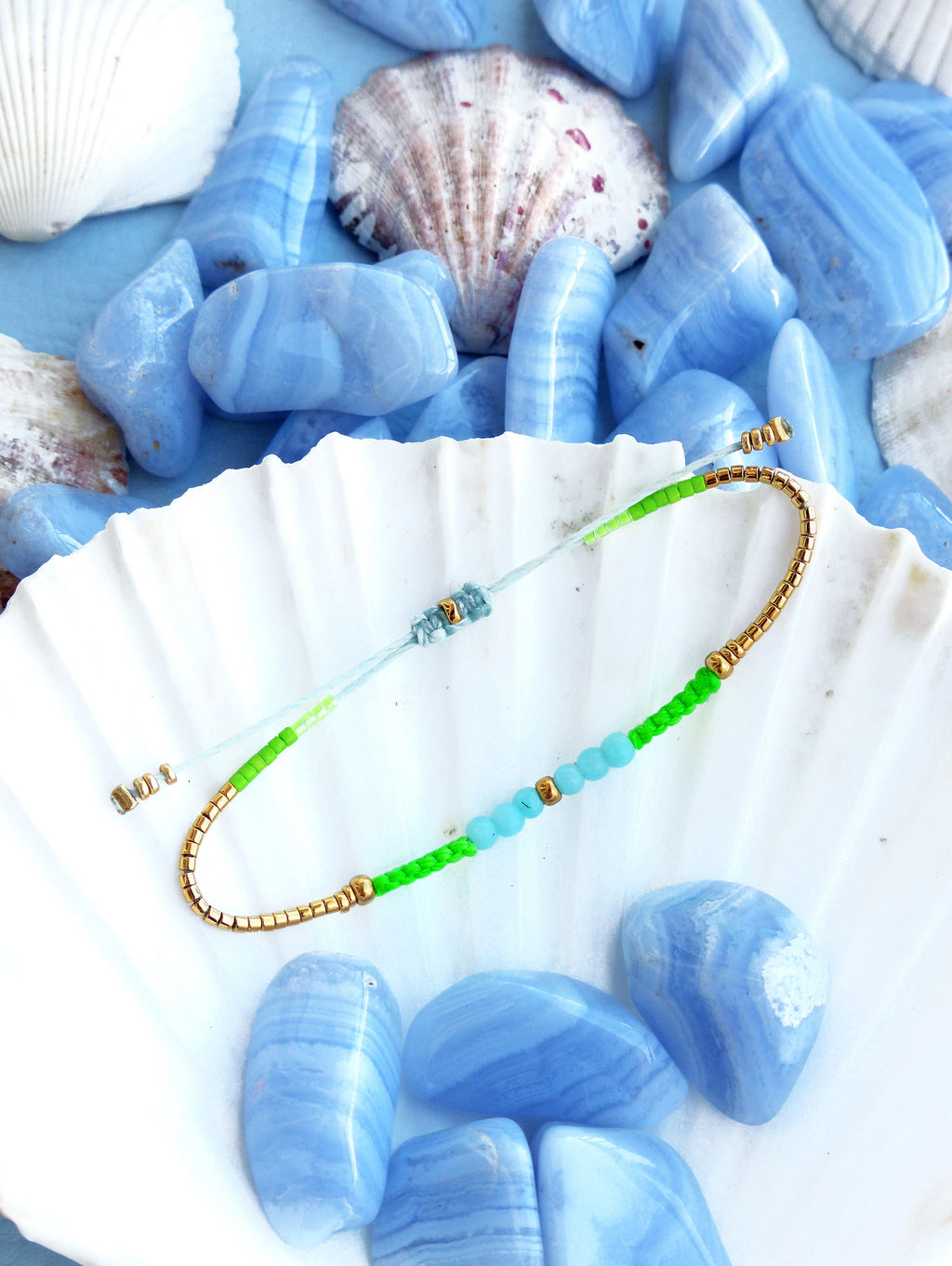 Peut inclure: Un bracelet délicat avec des perles vertes et bleues et des accents dorés. Le bracelet est présenté sur une coquille de mer blanche avec des pierres d'agate bleues.