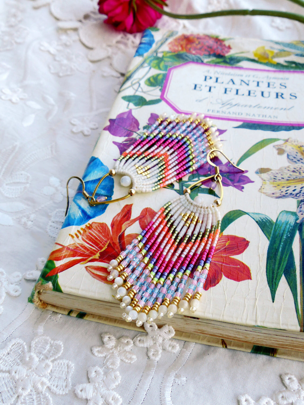 Peut inclure: Une paire de boucles d'oreilles en perles colorées avec un design en frange. Les boucles d'oreilles sont faites de perles blanches, roses, bleues, vertes et dorées. Les boucles d'oreilles sont accrochées à un livre avec un motif floral.