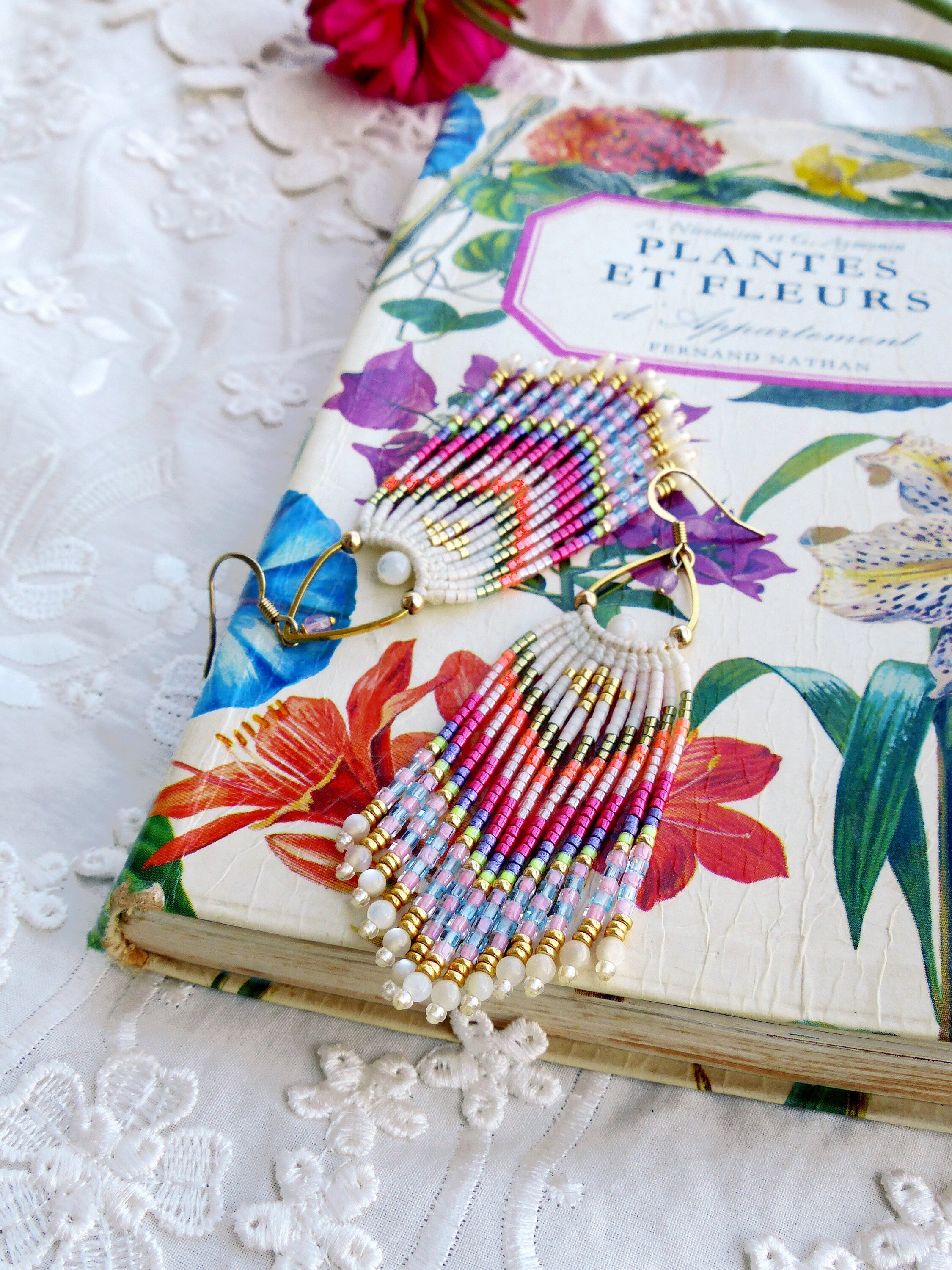 Peut inclure: Une paire de boucles d'oreilles en perles colorées avec un design en frange. Les boucles d'oreilles sont faites de perles blanches, roses, bleues, vertes et dorées. Les boucles d'oreilles sont accrochées à un livre avec un motif floral.
