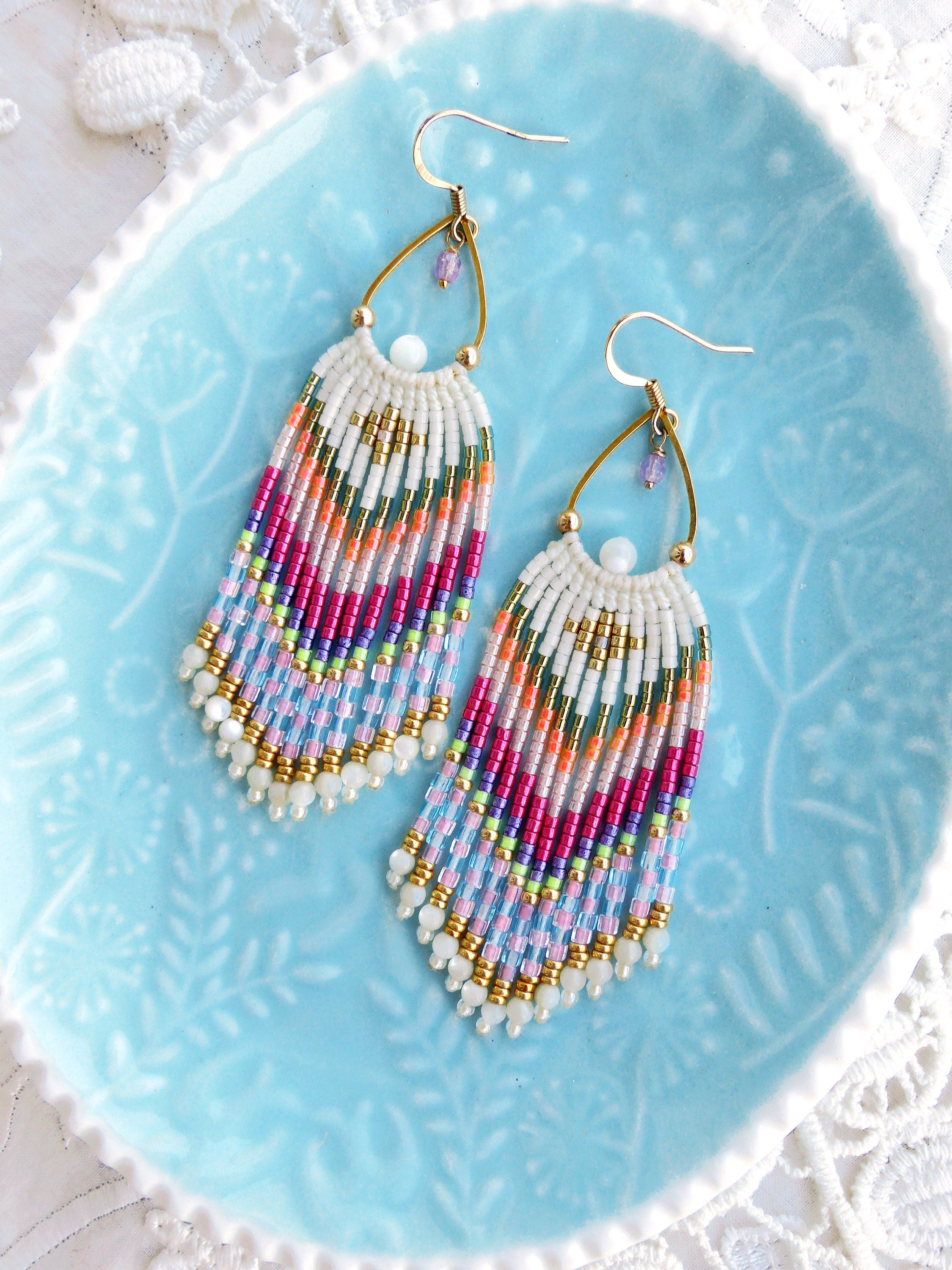 Peut inclure: Une paire de boucles d'oreilles pendantes en perles colorées avec un crochet en métal doré et une perle blanche en haut. Les boucles d'oreilles présentent une variété de couleurs, notamment le rose, le bleu, le vert et l'orange.