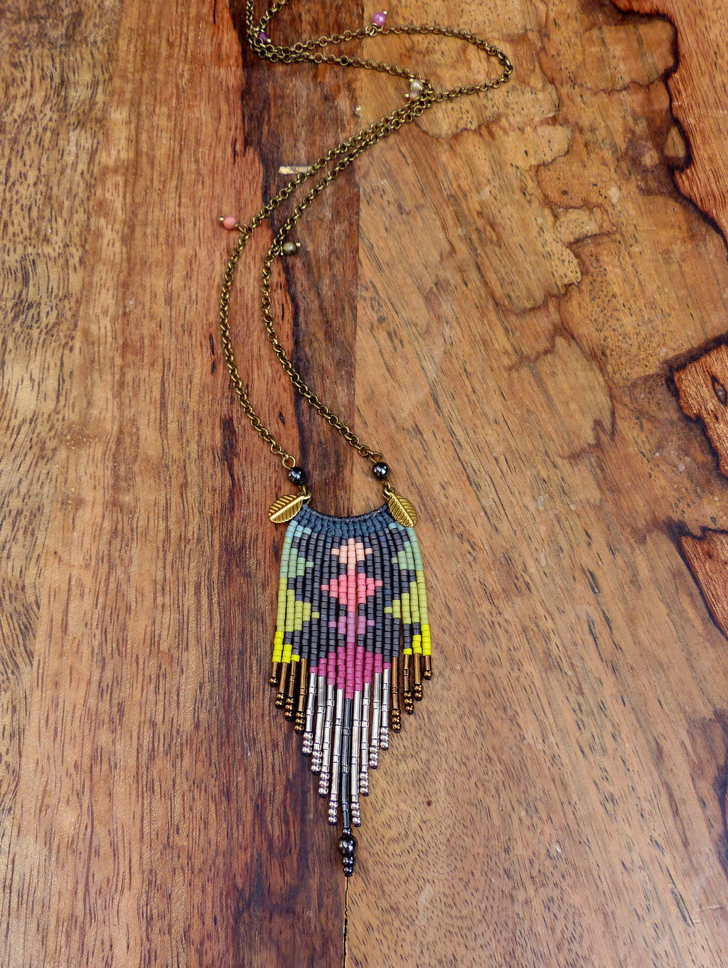 Peut inclure: Un long collier de perles avec un motif géométrique dans des tons de rose, vert, jaune et gris. Le collier a une frange en forme de glands en bas et est fait avec une variété de perles, y compris des perles métalliques.