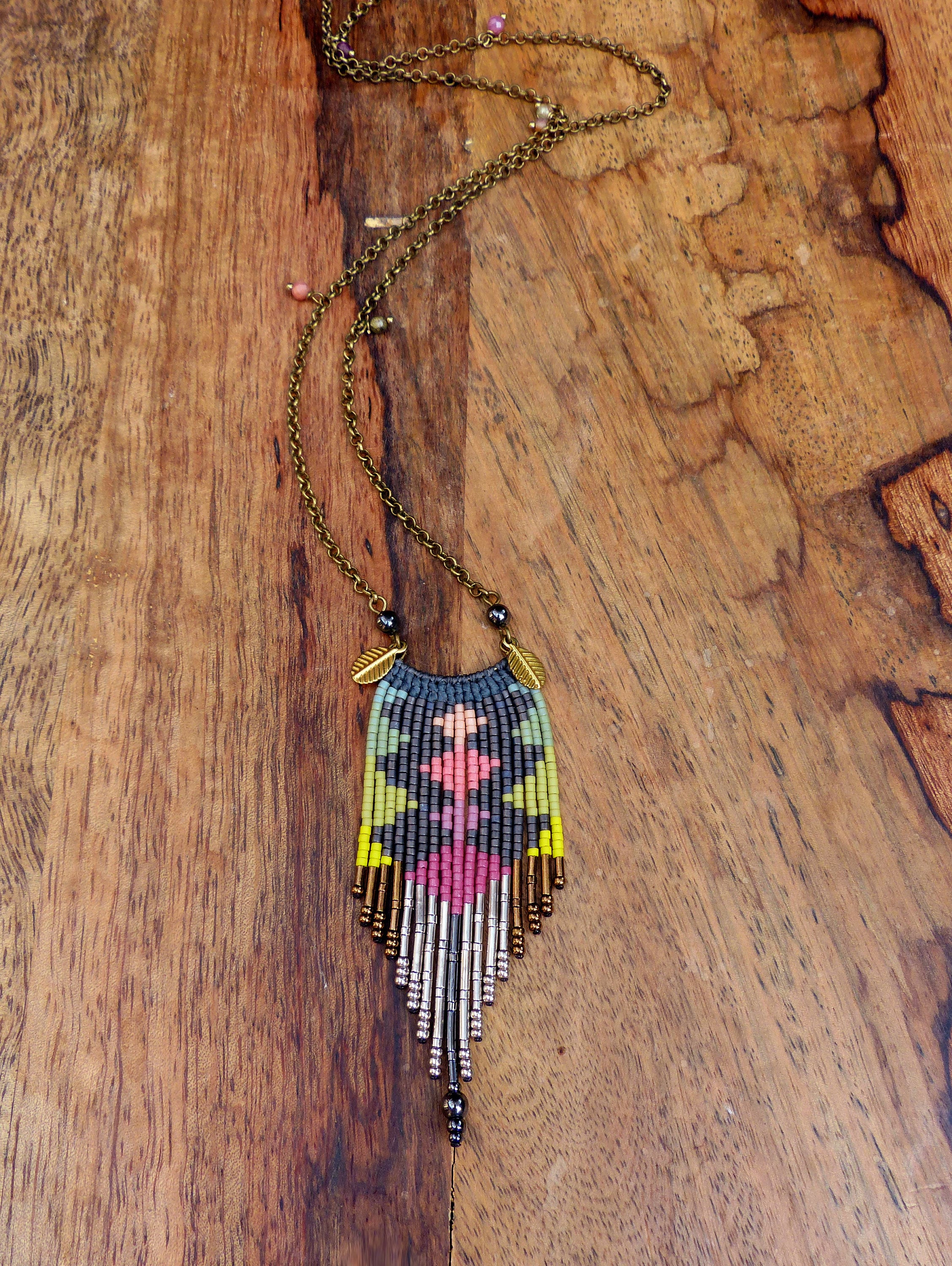 Peut inclure: Un long collier de perles avec un motif géométrique dans des tons de rose, vert, jaune et gris. Le collier a une frange en forme de glands en bas et est fait avec une variété de perles, y compris des perles métalliques.