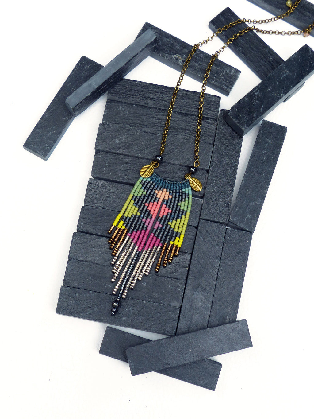 Peut inclure: Un collier de perles coloré avec un motif géométrique et des accents de feuilles d'or. Le collier est présenté sur un fond de schiste noir.