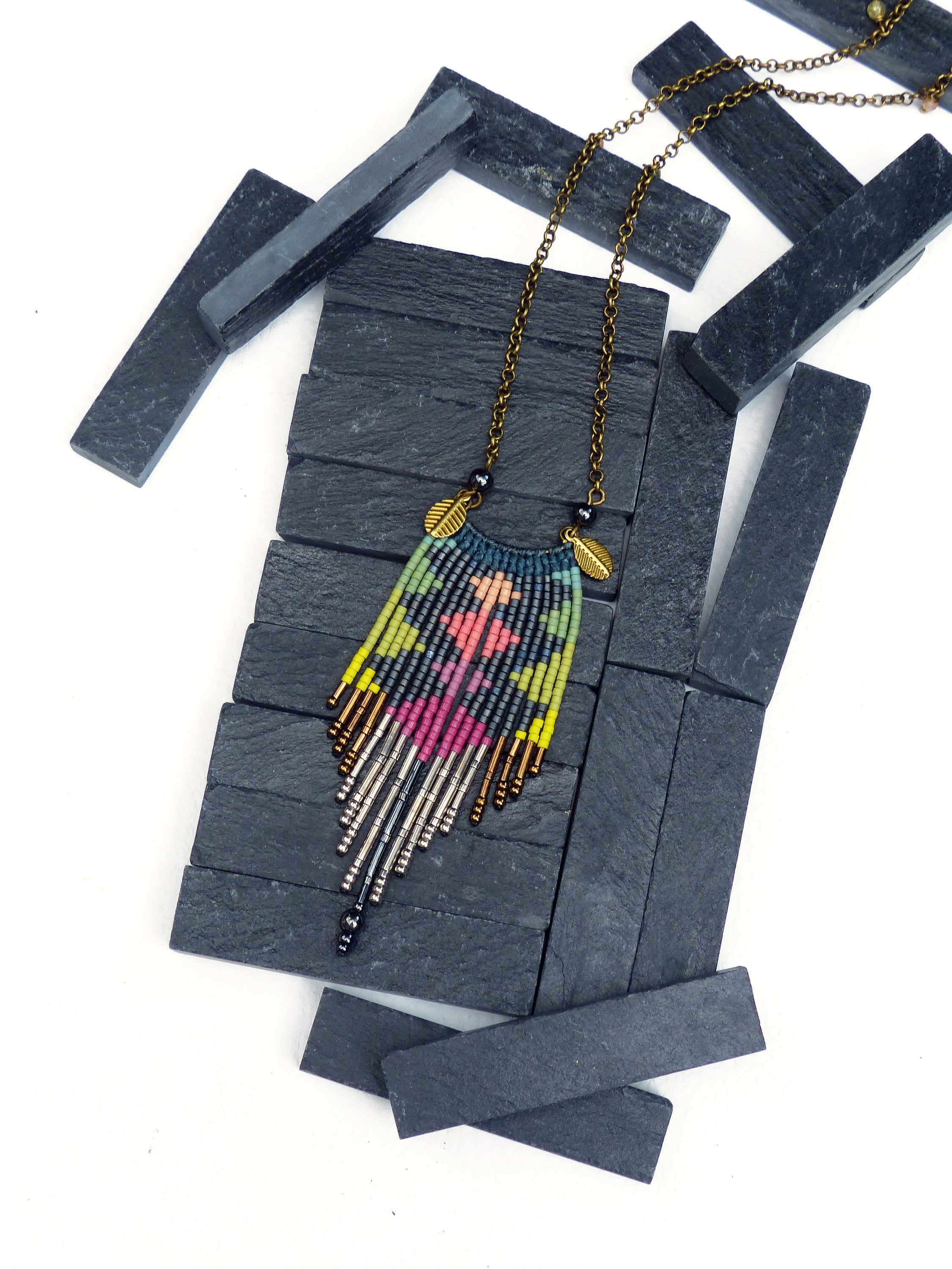 Peut inclure: Un collier de perles coloré avec un motif géométrique et des accents de feuilles d'or. Le collier est présenté sur un fond de schiste noir.