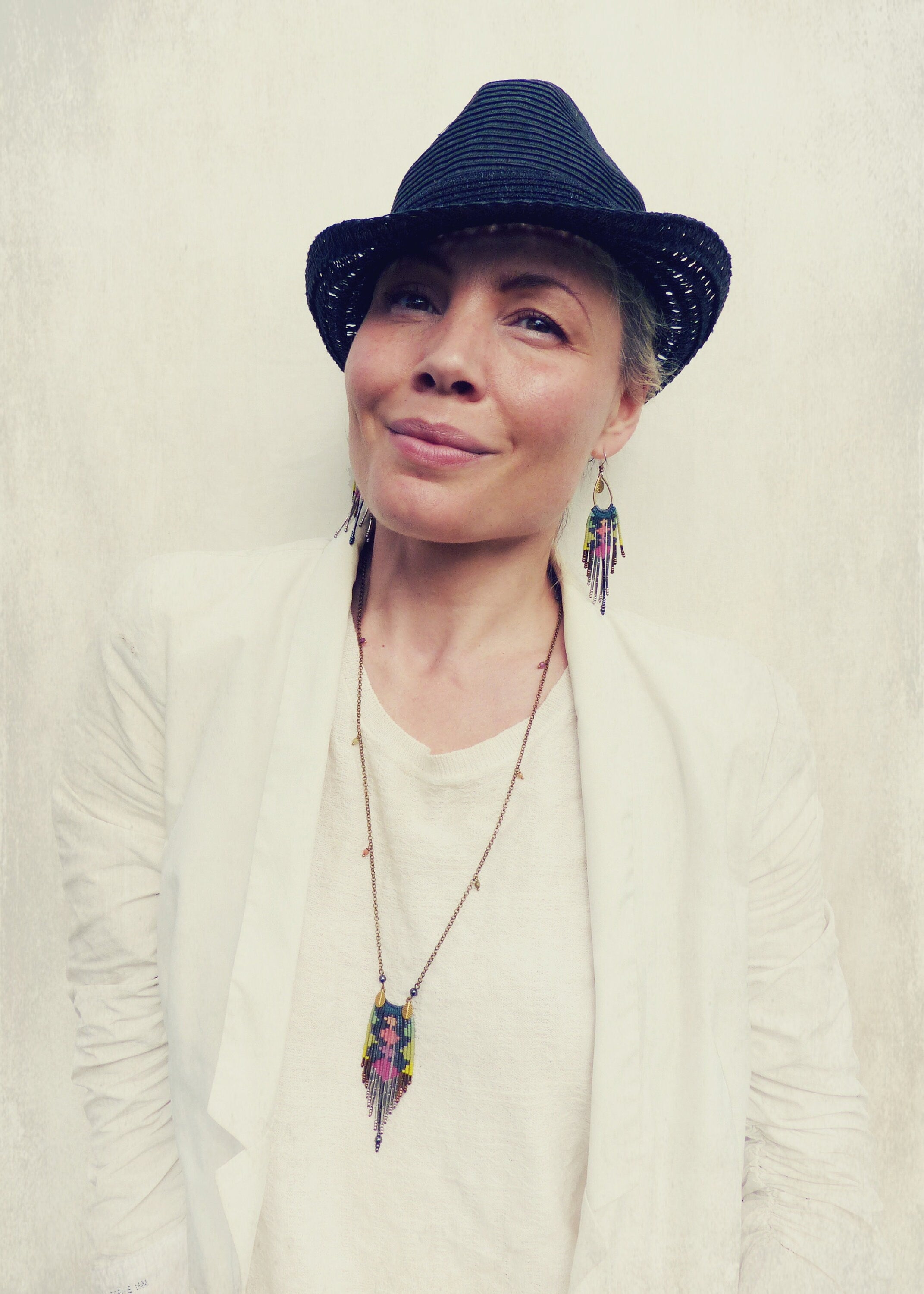 Peut inclure: Une femme portant un blazer blanc, un fedora noir, un long collier avec un pendentif coloré et des boucles d'oreilles à franges colorées.
