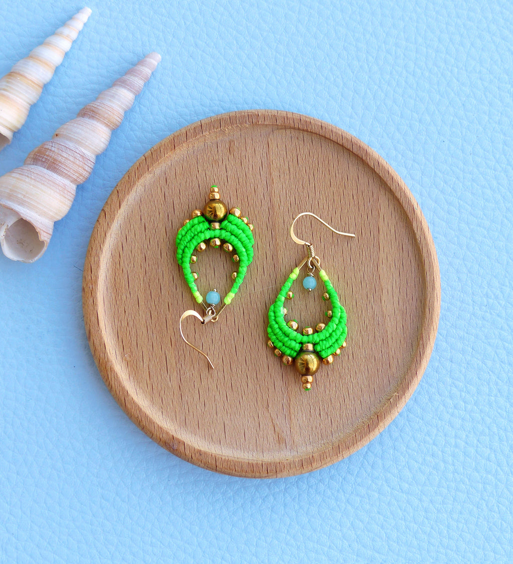 Peut inclure: Une paire de boucles d'oreilles en forme de larme, composées de perles vertes et dorées. Les boucles d'oreilles sont posées sur une surface en bois.