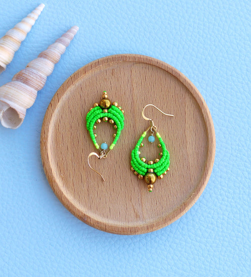 Peut inclure: Une paire de boucles d'oreilles en forme de larme, composées de perles vertes et dorées. Les boucles d'oreilles sont posées sur une surface en bois.