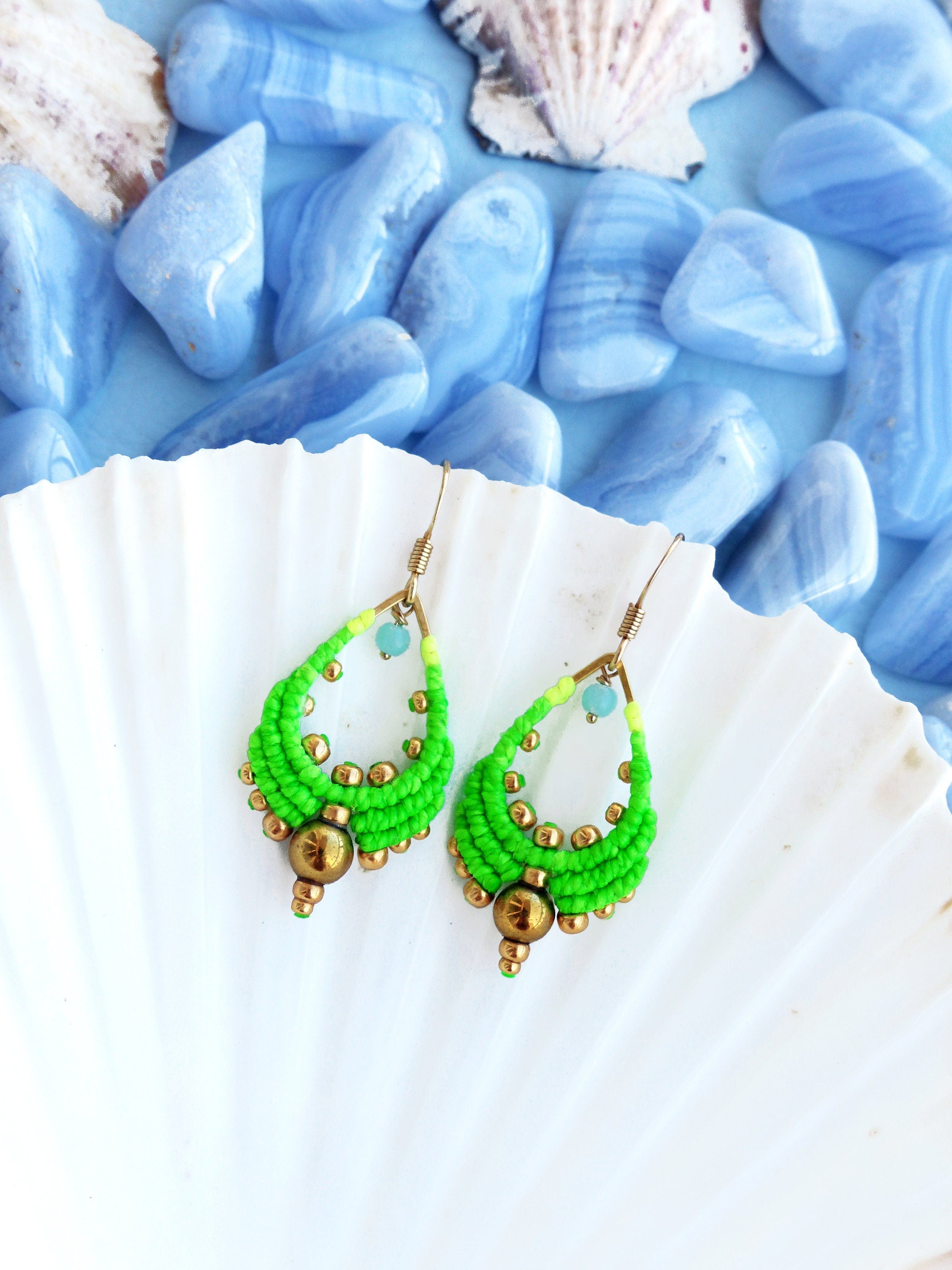 Peut inclure: Une paire de boucles d'oreilles en forme de larme vertes et dorées avec un design perlé. Les boucles d'oreilles sont sur un fond de coquille de mer blanche.