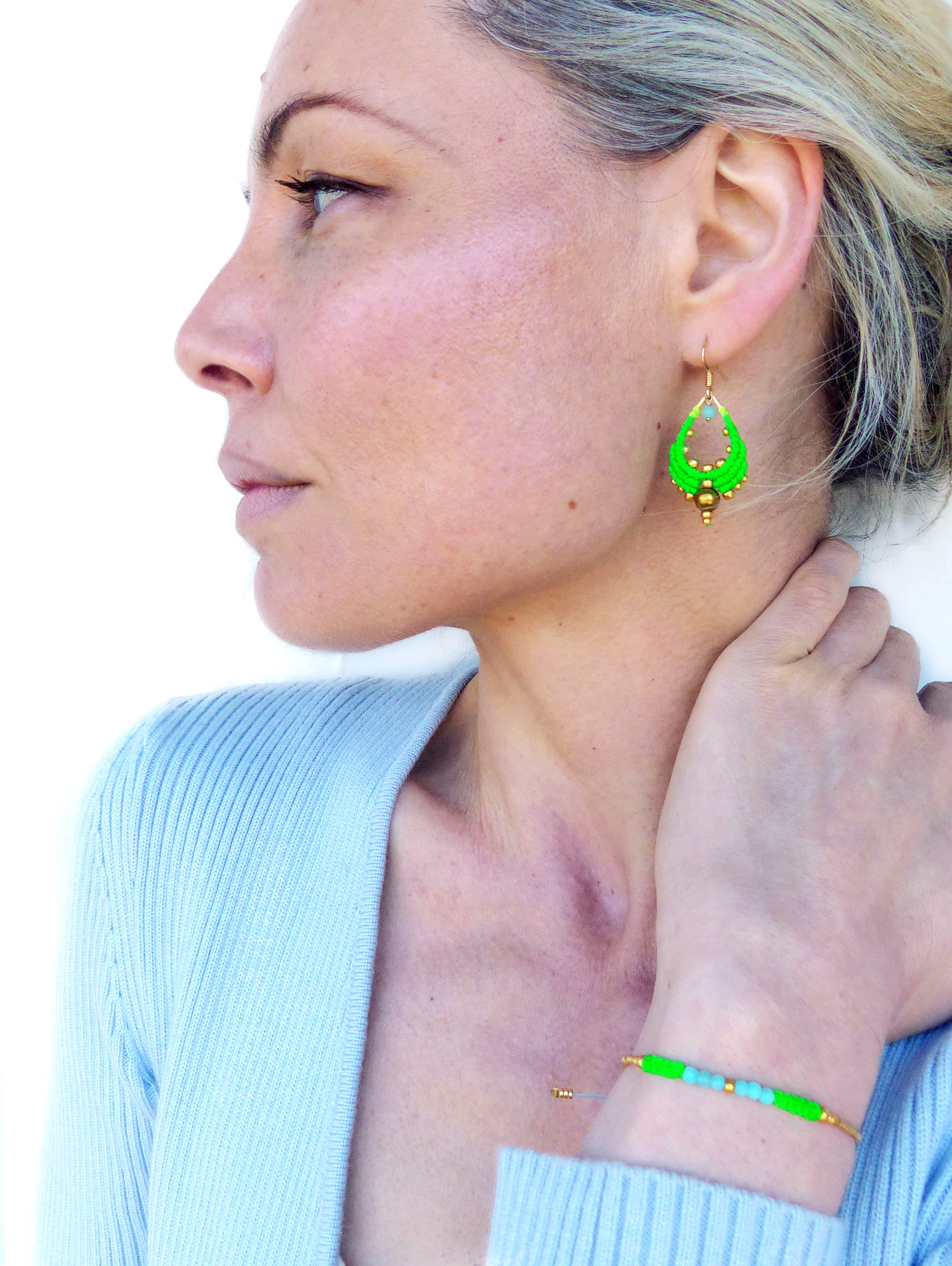 Peut inclure: Une femme portant un pull bleu clair avec un bracelet en perles vertes et dorées au poignet. Elle porte également une paire de boucles d'oreilles pendantes vertes et dorées.