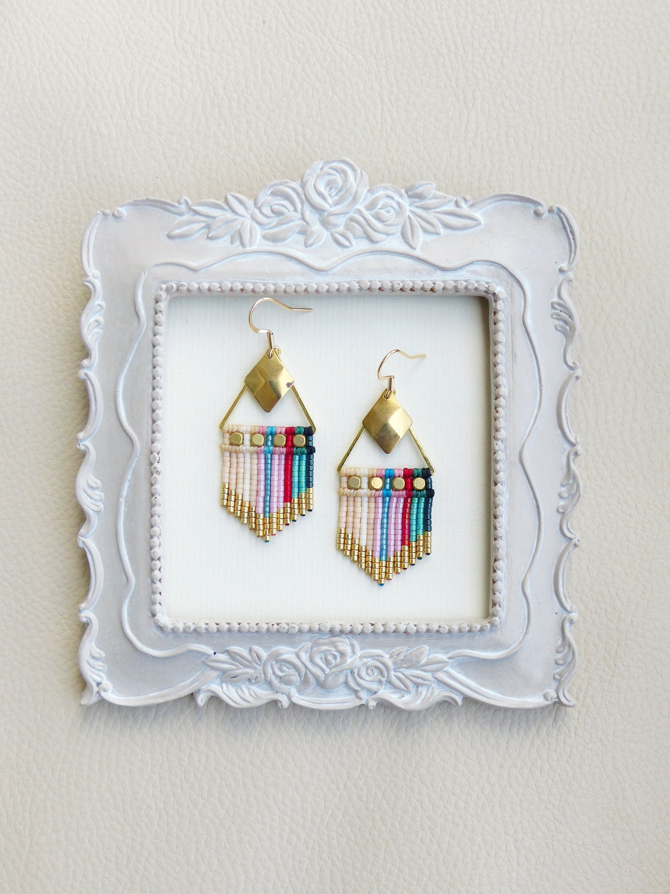 Peut inclure: Une paire de boucles d'oreilles en perles colorées avec des accents dorés. Les boucles d'oreilles sont présentées dans un cadre blanc à motif floral.