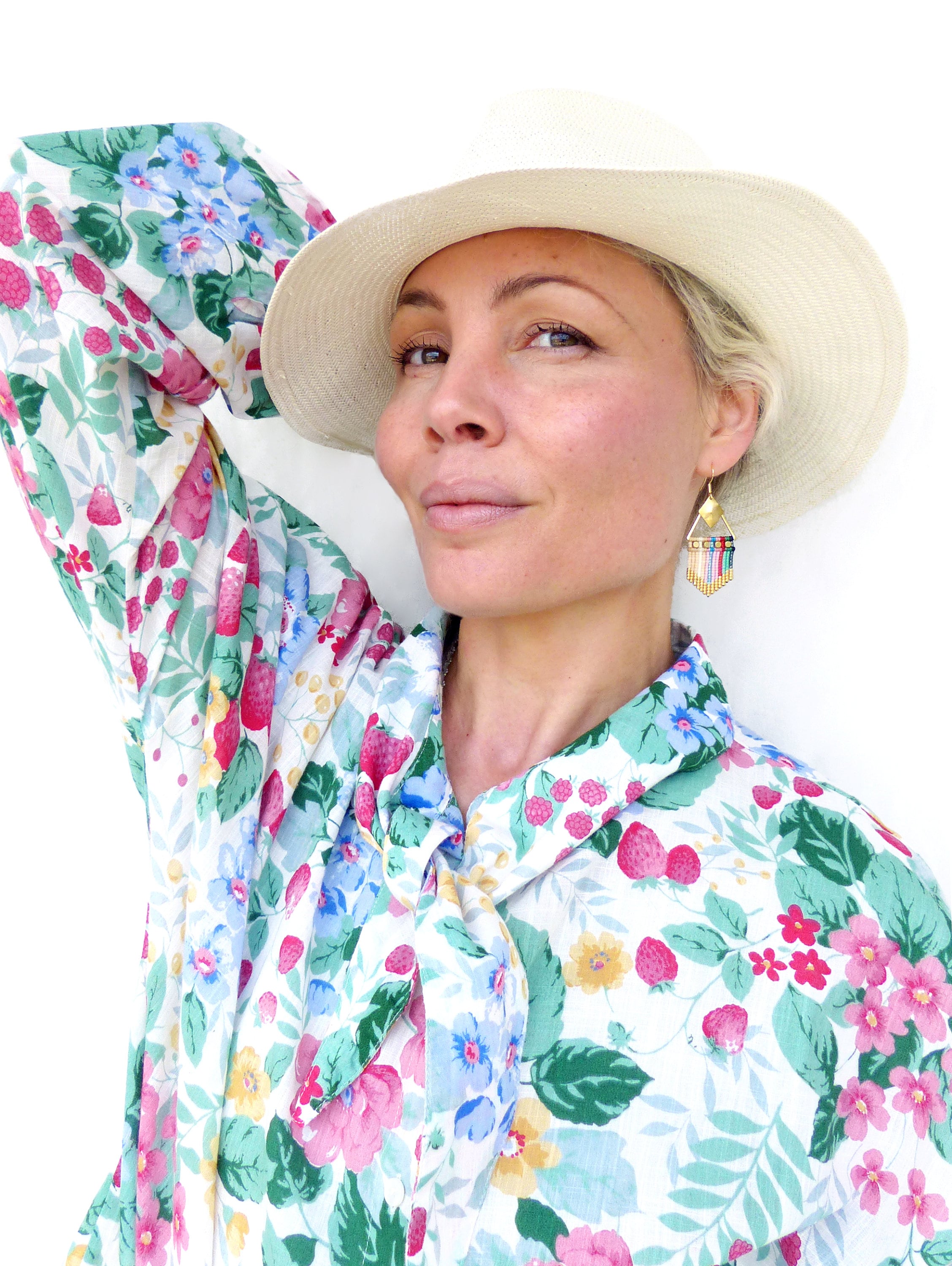 Peut inclure: Une femme aux cheveux blonds porte un chapeau blanc à large bord et une blouse à imprimé floral. Elle porte une paire de boucles d'oreilles en or avec des perles colorées.