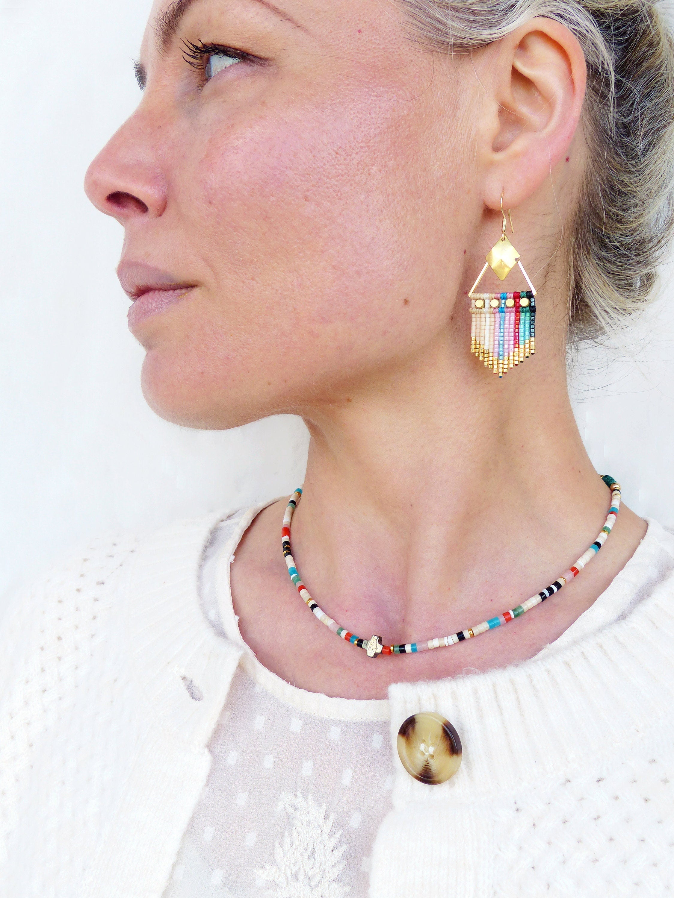 Peut inclure: Une femme portant un cardigan blanc et une chemise blanche à petits pois. Elle porte un collier de perles colorées et des boucles d'oreilles assorties. Les boucles d'oreilles sont en or et ont une forme géométrique avec une frange de perles.