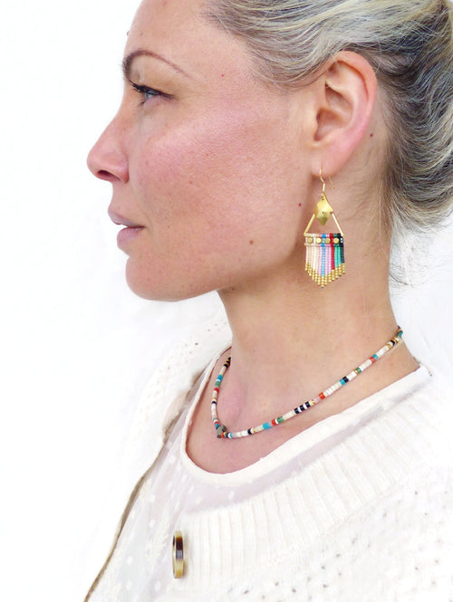 Peut inclure: Une femme portant un pull blanc et un collier de perles colorées. Elle porte également des boucles d'oreilles en or avec un motif géométrique et des perles colorées.
