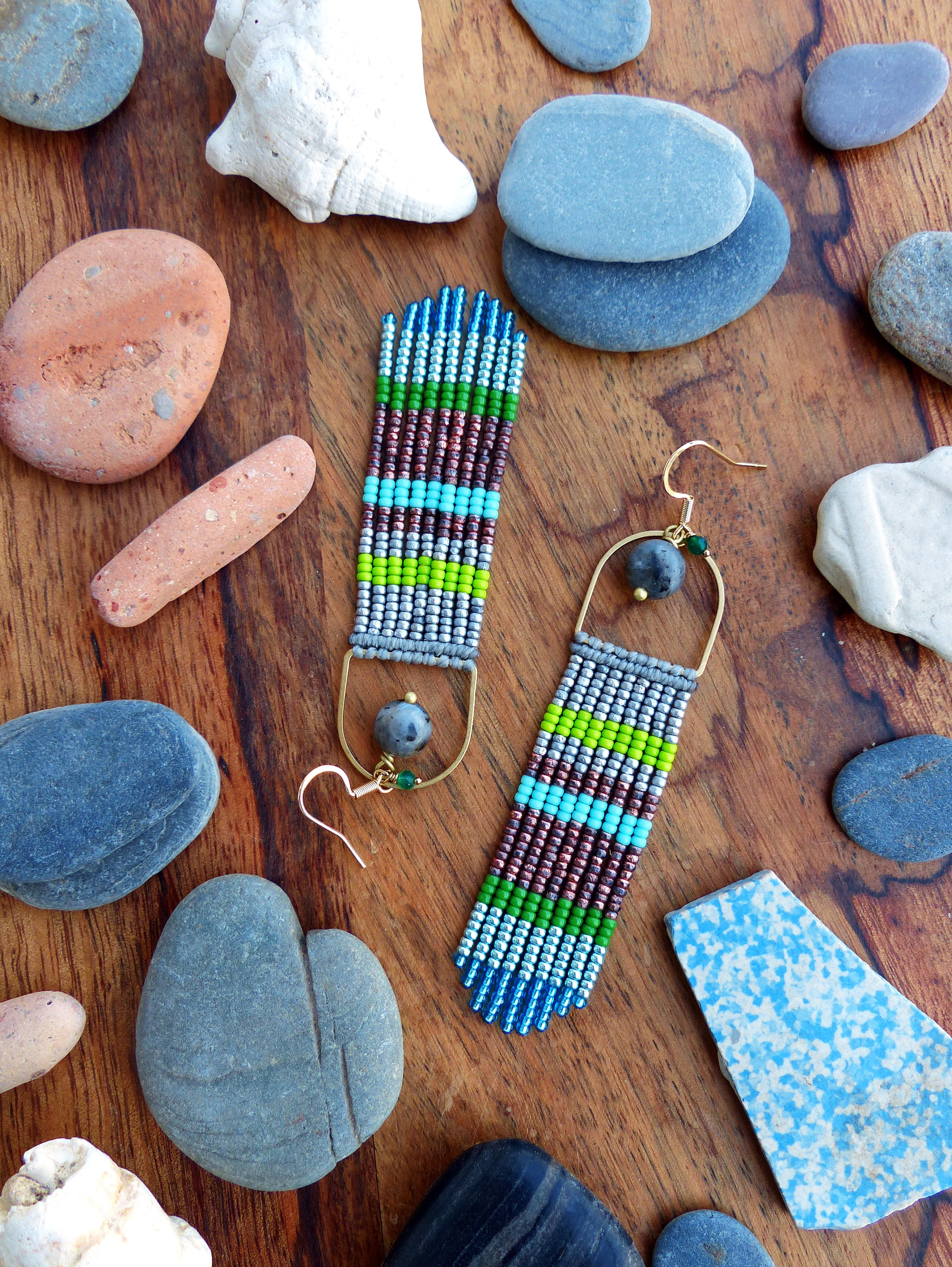 Peut inclure: Une paire de longues boucles d'oreilles en perles avec un schéma de couleurs argent, vert, bleu et marron. Les boucles d'oreilles sont suspendues à des crochets dorés.