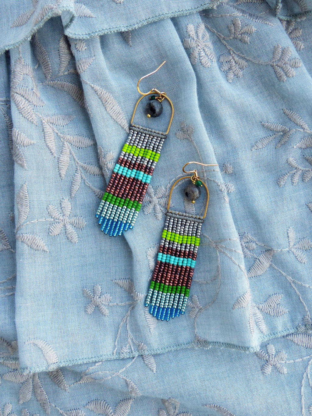 Peut inclure: Une paire de longues boucles d'oreilles en perles avec un motif géométrique. Les boucles d'oreilles sont faites de différentes couleurs, notamment le bleu, le vert, le gris et le marron. Les boucles d'oreilles sont suspendues à des créoles dorées.
