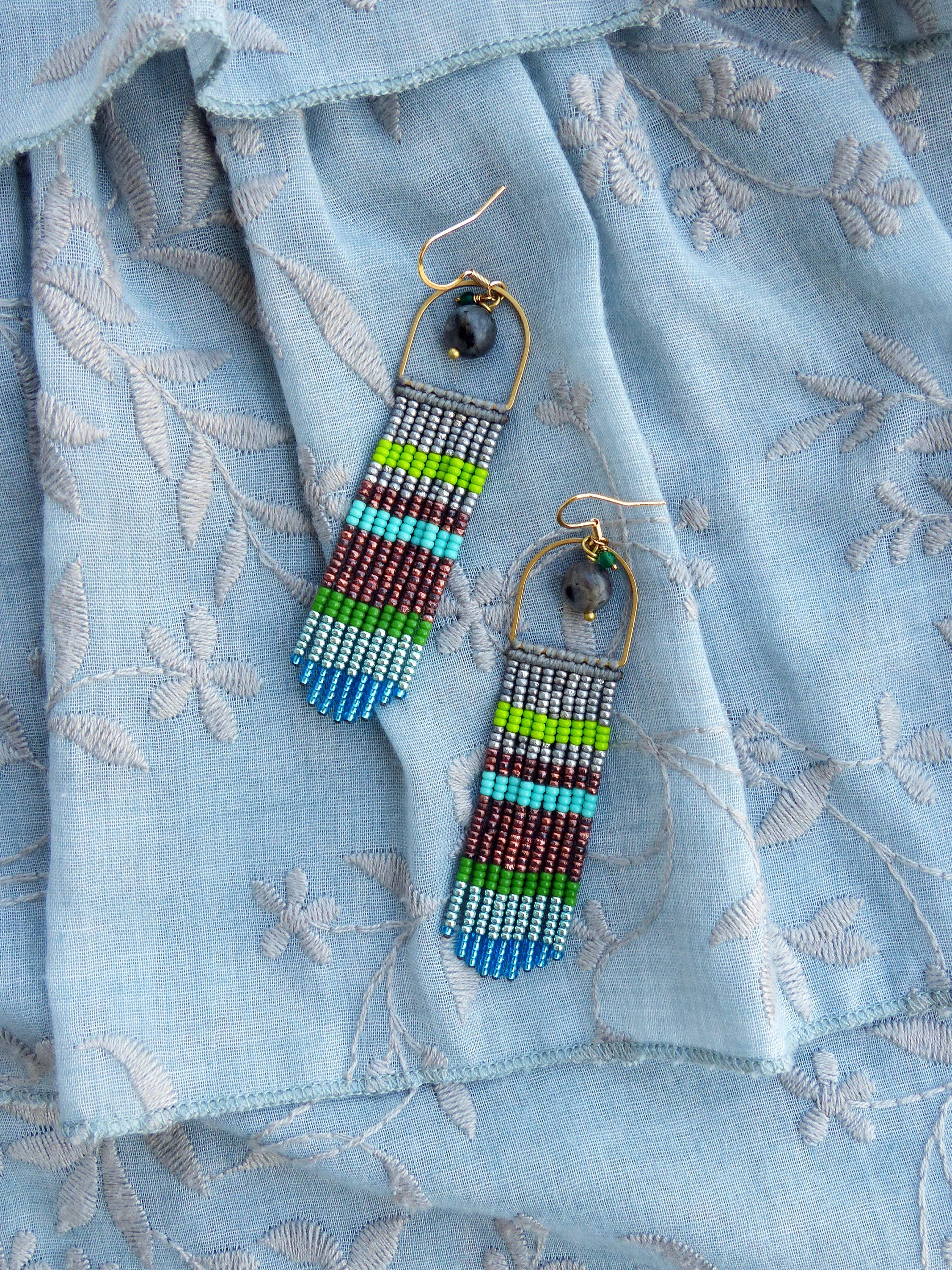 Peut inclure: Une paire de longues boucles d'oreilles en perles avec un motif géométrique. Les boucles d'oreilles sont faites de différentes couleurs, notamment le bleu, le vert, le gris et le marron. Les boucles d'oreilles sont suspendues à des créoles dorées.