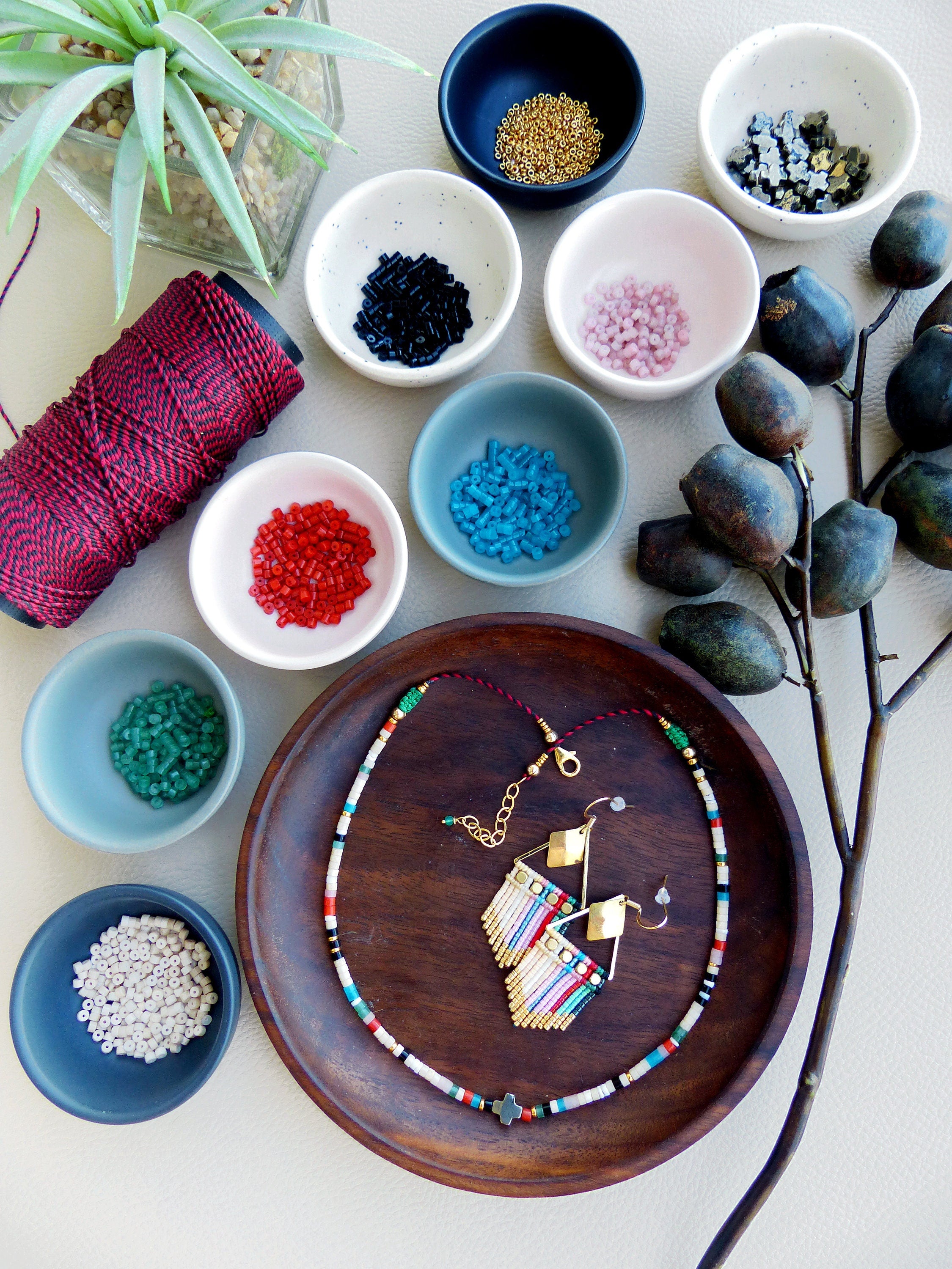 Peut inclure: Un collier et des boucles d'oreilles en perles colorées avec des accents dorés. Le collier est sur une assiette en bois avec de petits bols de perles de différentes couleurs autour.
