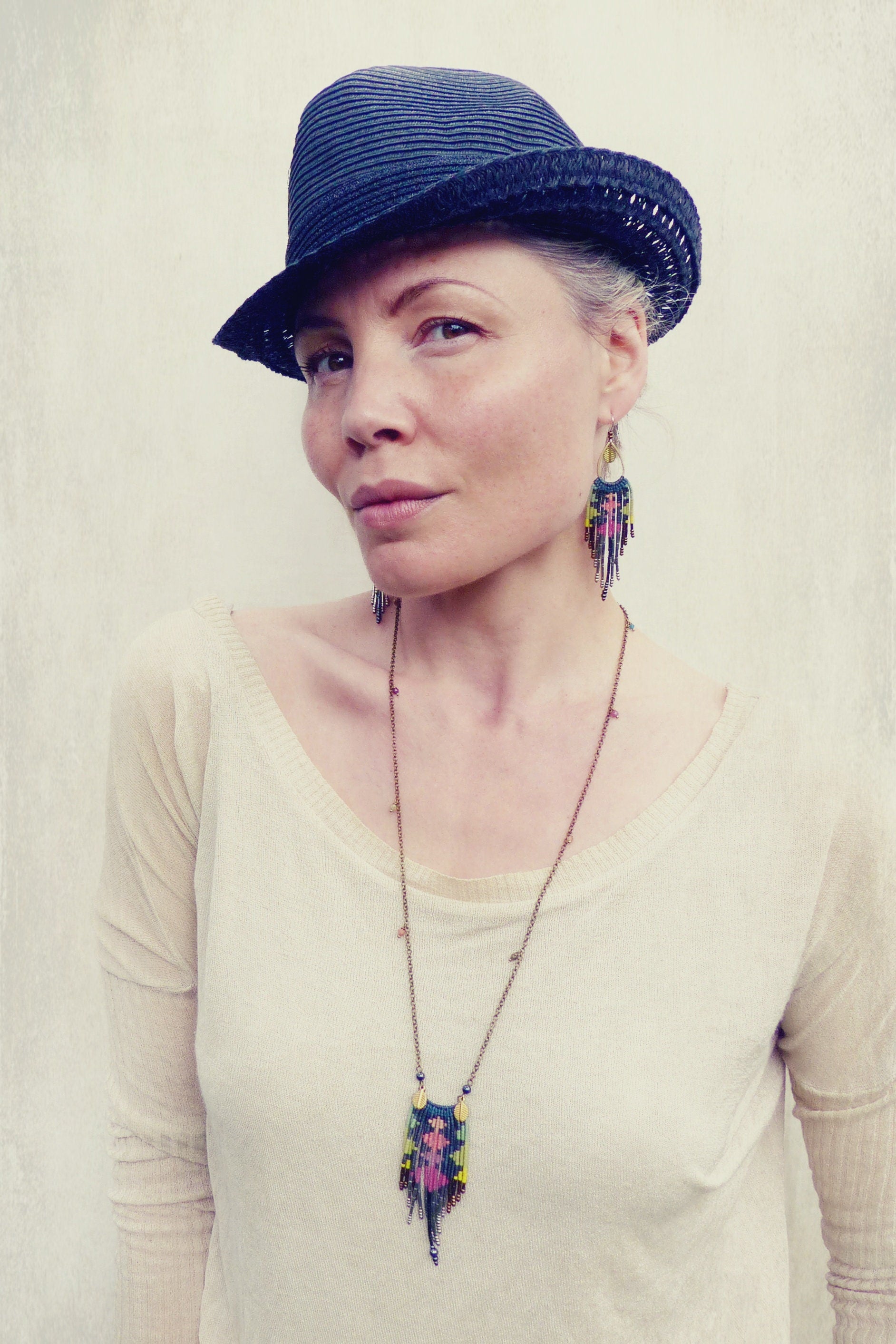 Peut inclure: Une femme portant un chapeau fedora noir, un pull blanc et un collier de perles avec un pendentif coloré. Elle porte également des boucles d'oreilles en perles.