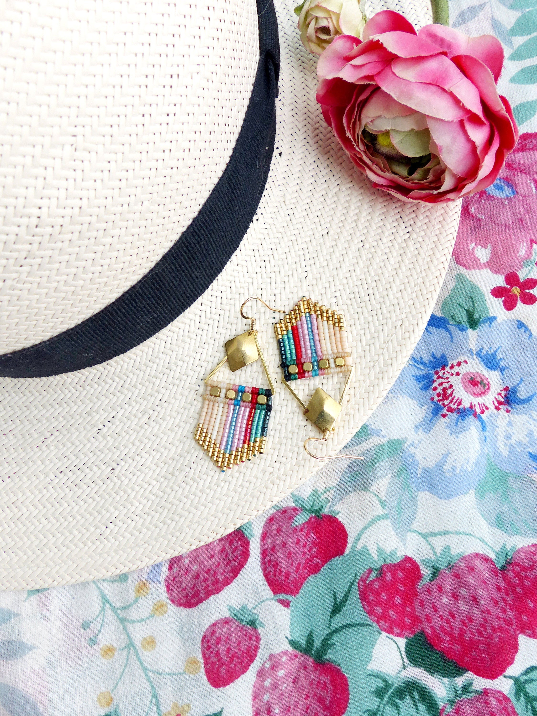 Peut inclure: Une paire de boucles d'oreilles en perles colorées avec des accents dorés. Les boucles d'oreilles sont suspendues à un chapeau de paille et un tissu à motifs floraux.