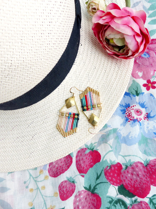 Peut inclure: Une paire de boucles d'oreilles en perles colorées avec des accents dorés. Les boucles d'oreilles sont suspendues à un chapeau de paille et un tissu à motifs floraux.