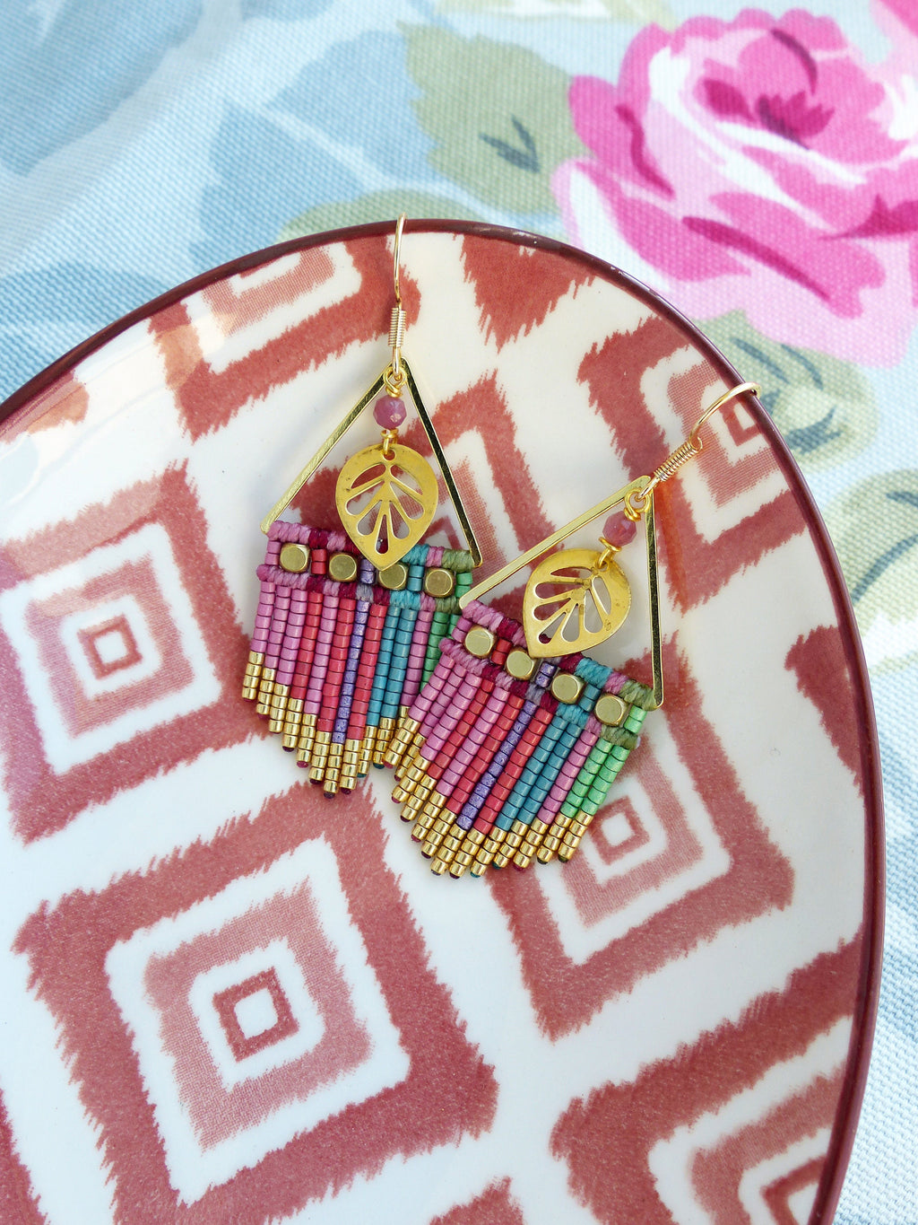 Peut inclure: Une paire de boucles d'oreilles dorées avec des franges de perles roses, vertes et bleues et un charme en forme de feuille. Les boucles d'oreilles sont sur une assiette blanche avec un motif géométrique rouge et blanc.