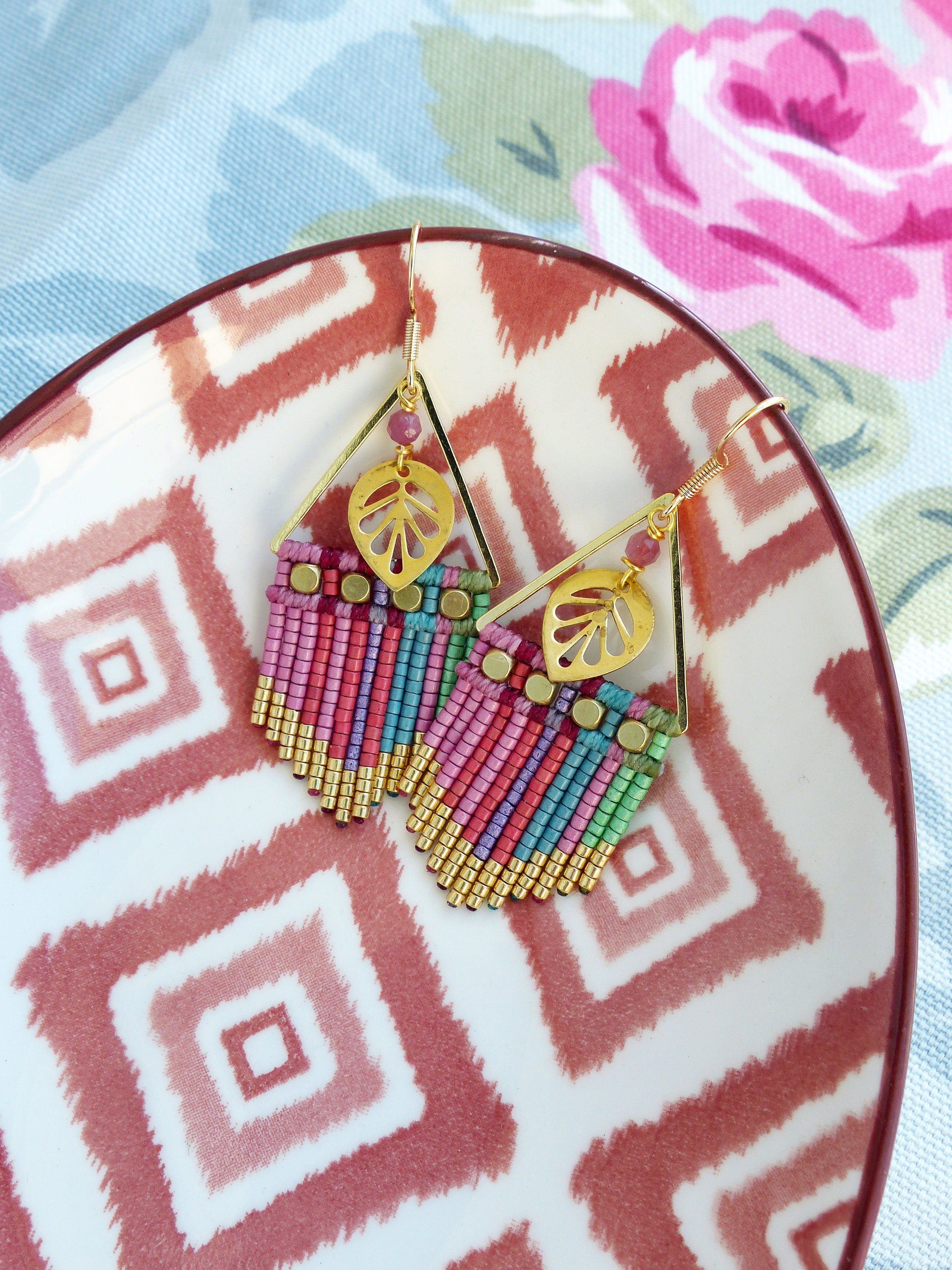 Peut inclure: Une paire de boucles d'oreilles dorées avec des franges de perles roses, vertes et bleues et un charme en forme de feuille. Les boucles d'oreilles sont sur une assiette blanche avec un motif géométrique rouge et blanc.