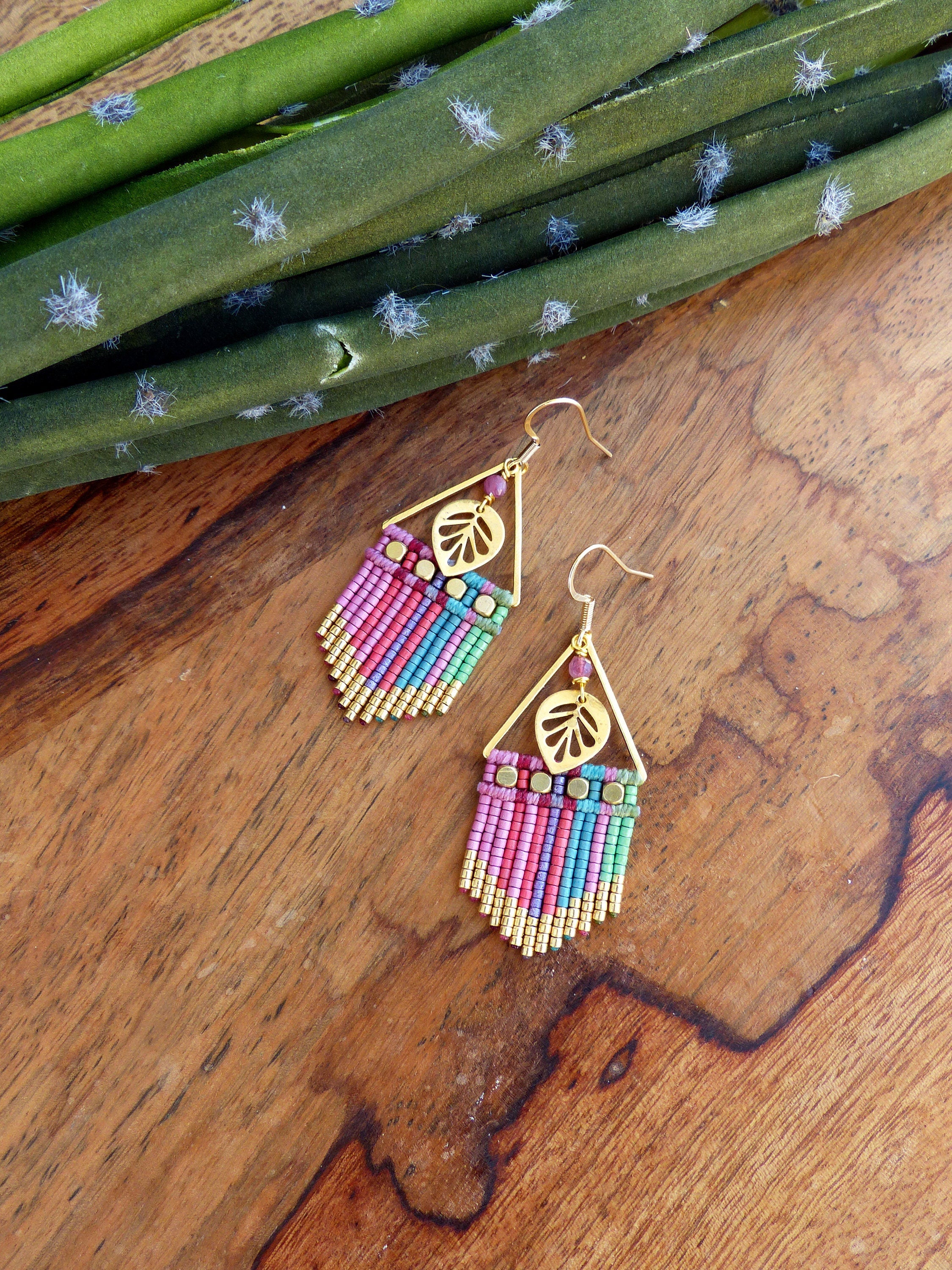 Peut inclure: Une paire de boucles d'oreilles pendantes dorées avec un motif de feuille et une frange de perles colorées. Les boucles d'oreilles sont sur une surface en bois.
