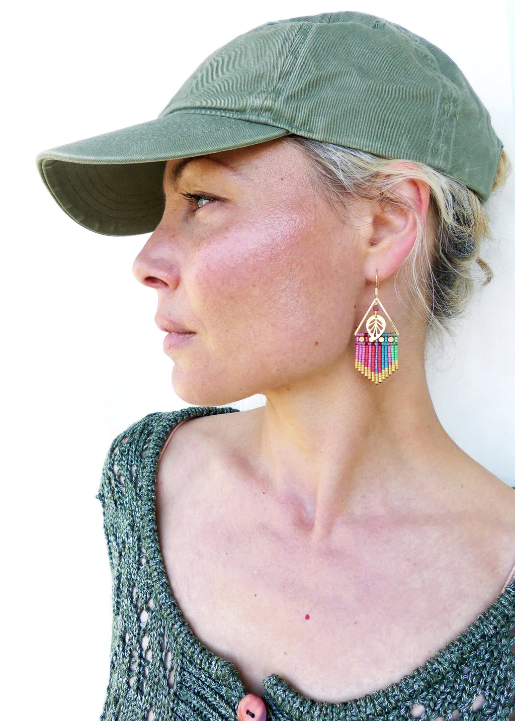 Peut inclure: Une femme portant un pull en tricot vert et une casquette de baseball verte. Elle porte des boucles d'oreilles en perles colorées avec un charme en forme de signe de paix.