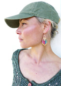 Peut inclure: Une femme portant un pull en tricot vert et une casquette de baseball verte. Elle porte des boucles d'oreilles en perles colorées avec un charme en forme de signe de paix.