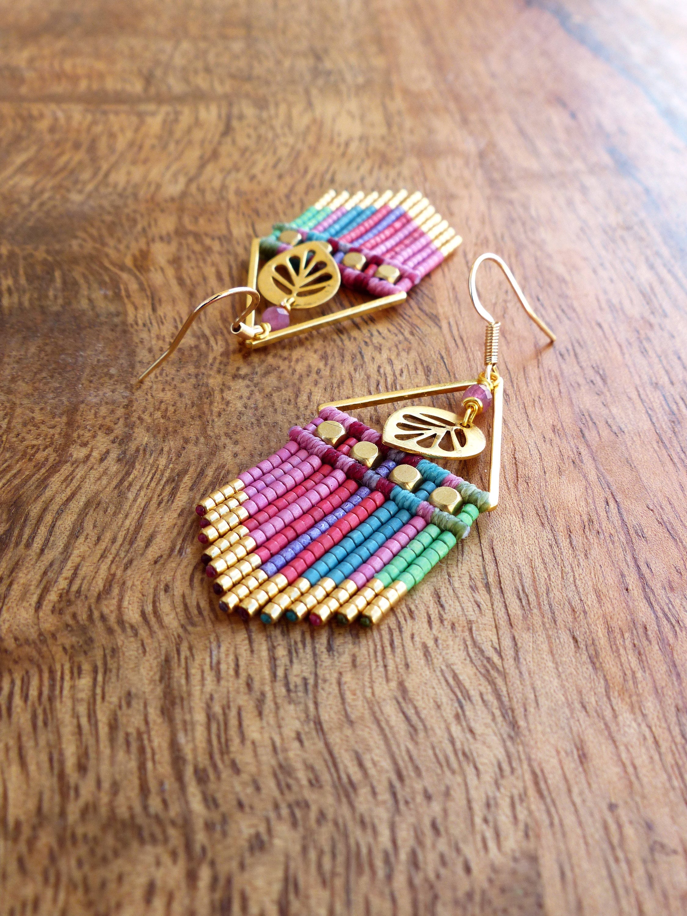 Peut inclure: Une paire de boucles d'oreilles en perles colorées avec des accents dorés et un charme en forme de feuille. Les boucles d'oreilles sont faites de petites perles colorées et ont une forme géométrique.