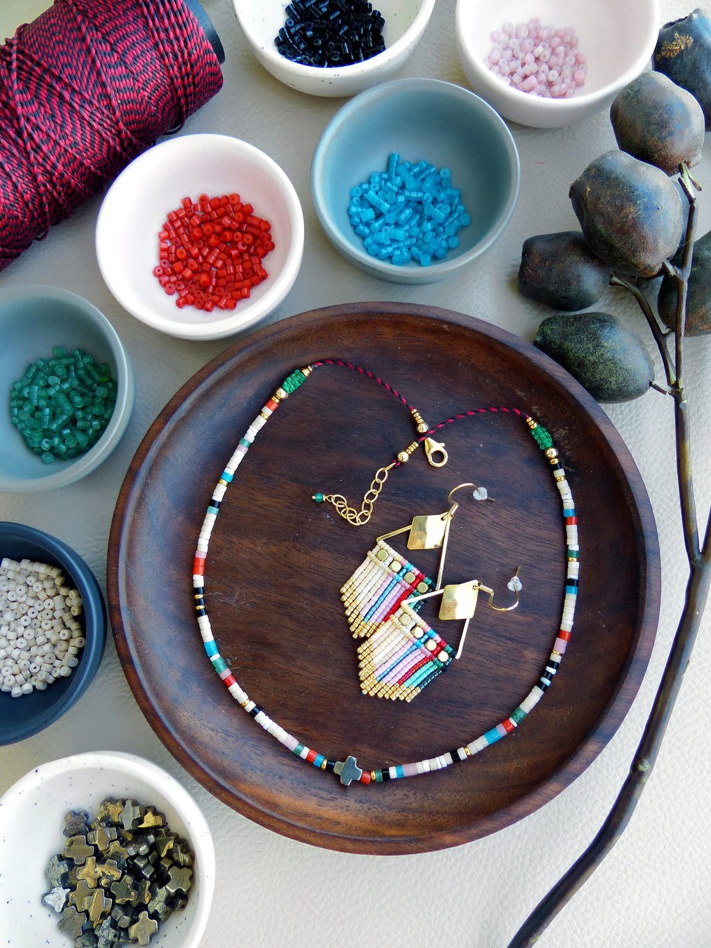 Peut inclure: Un collier et des boucles d'oreilles en perles colorées avec un pendentif en forme de croix dorée. Le collier et les boucles d'oreilles sont disposés sur une assiette en bois avec diverses perles dans de petits bols autour.