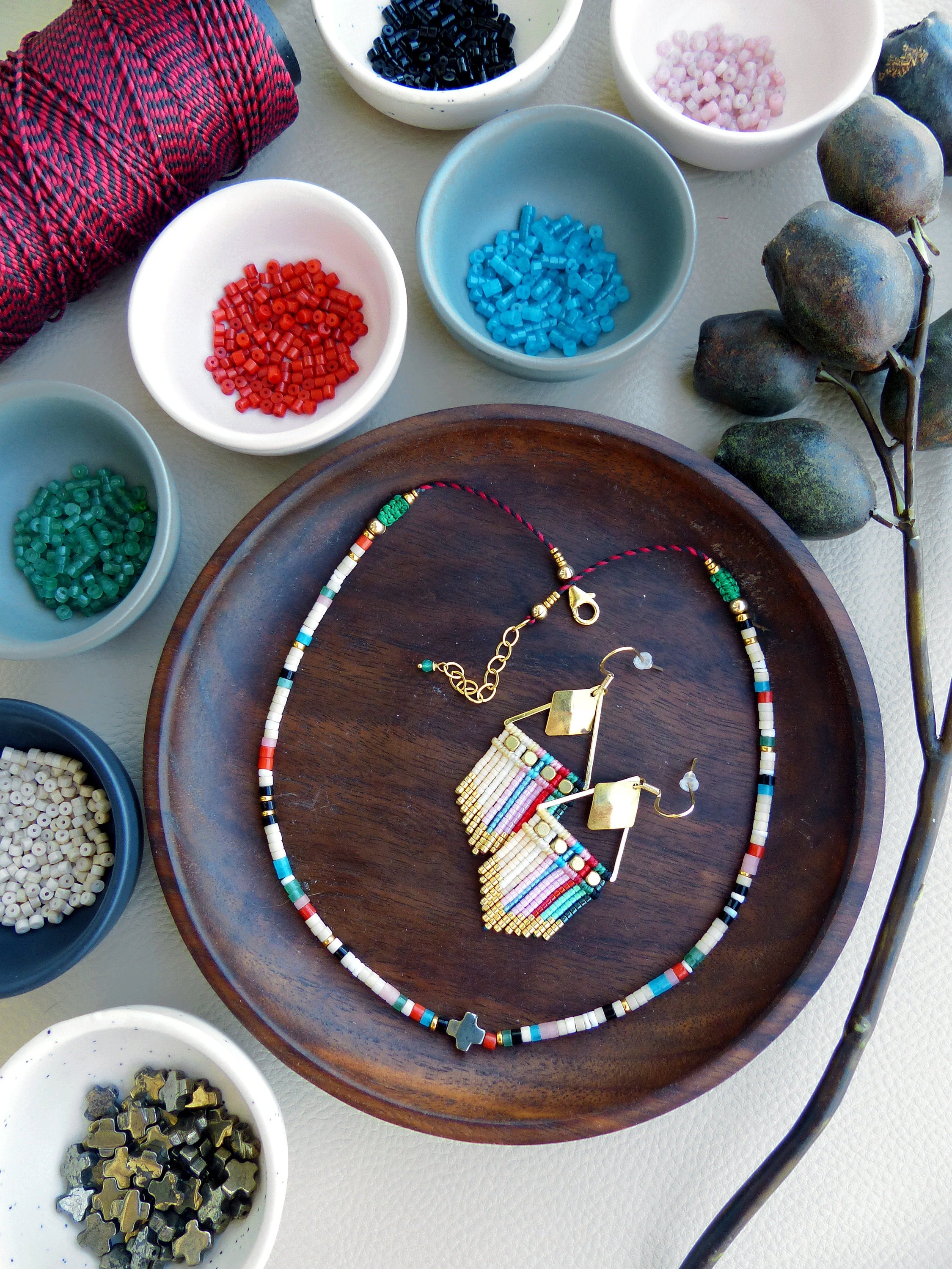 Peut inclure: Un collier et des boucles d'oreilles en perles colorées avec un pendentif en forme de croix dorée. Le collier et les boucles d'oreilles sont disposés sur une assiette en bois avec diverses perles dans de petits bols autour.