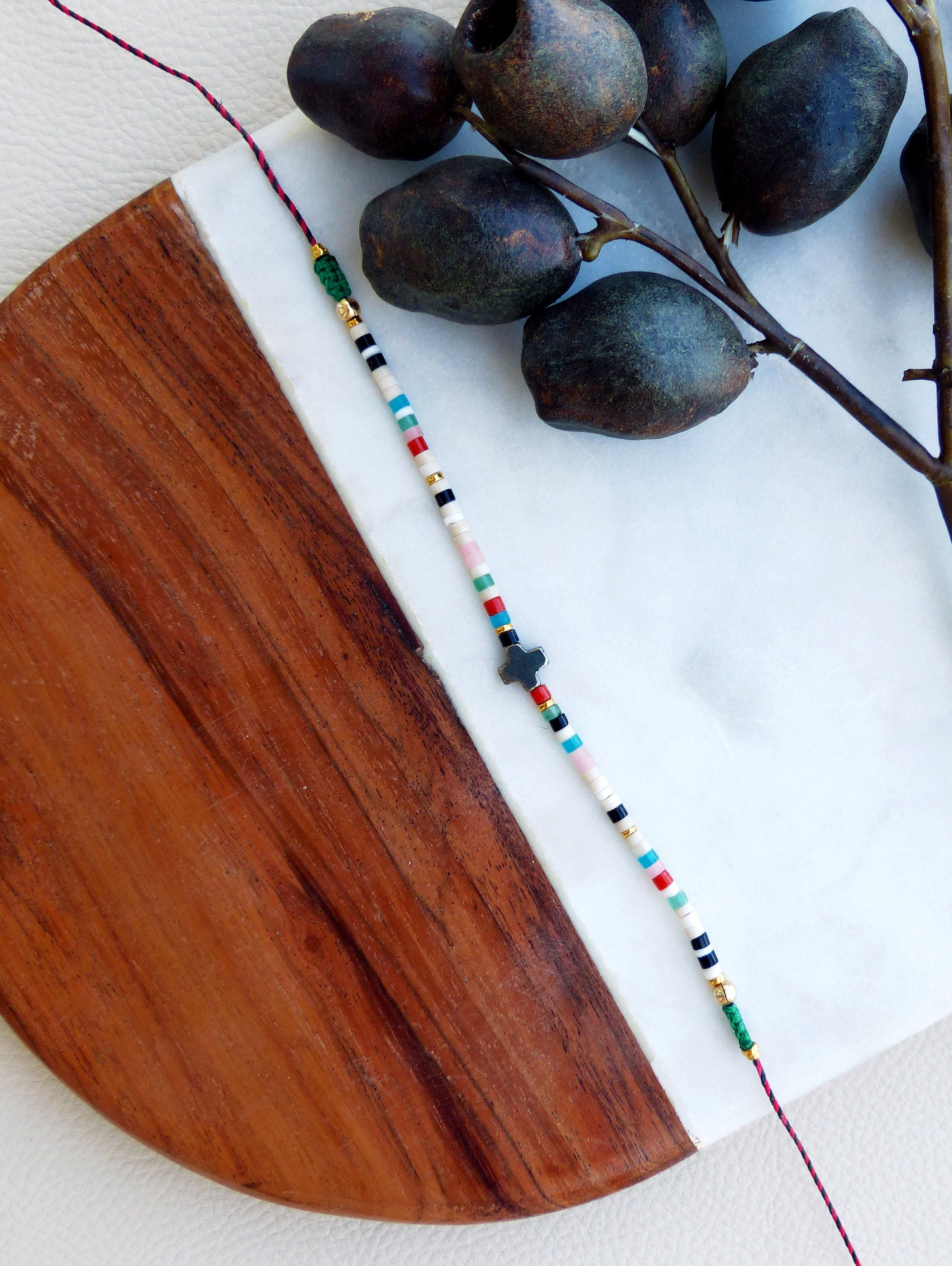 Peut inclure: Un bracelet en perles colorées avec un pendentif en forme de croix noire. Le bracelet est composé de perles blanches, noires, rouges, bleues, vertes et dorées. Le bracelet est posé sur une surface blanche avec un fond en bois.